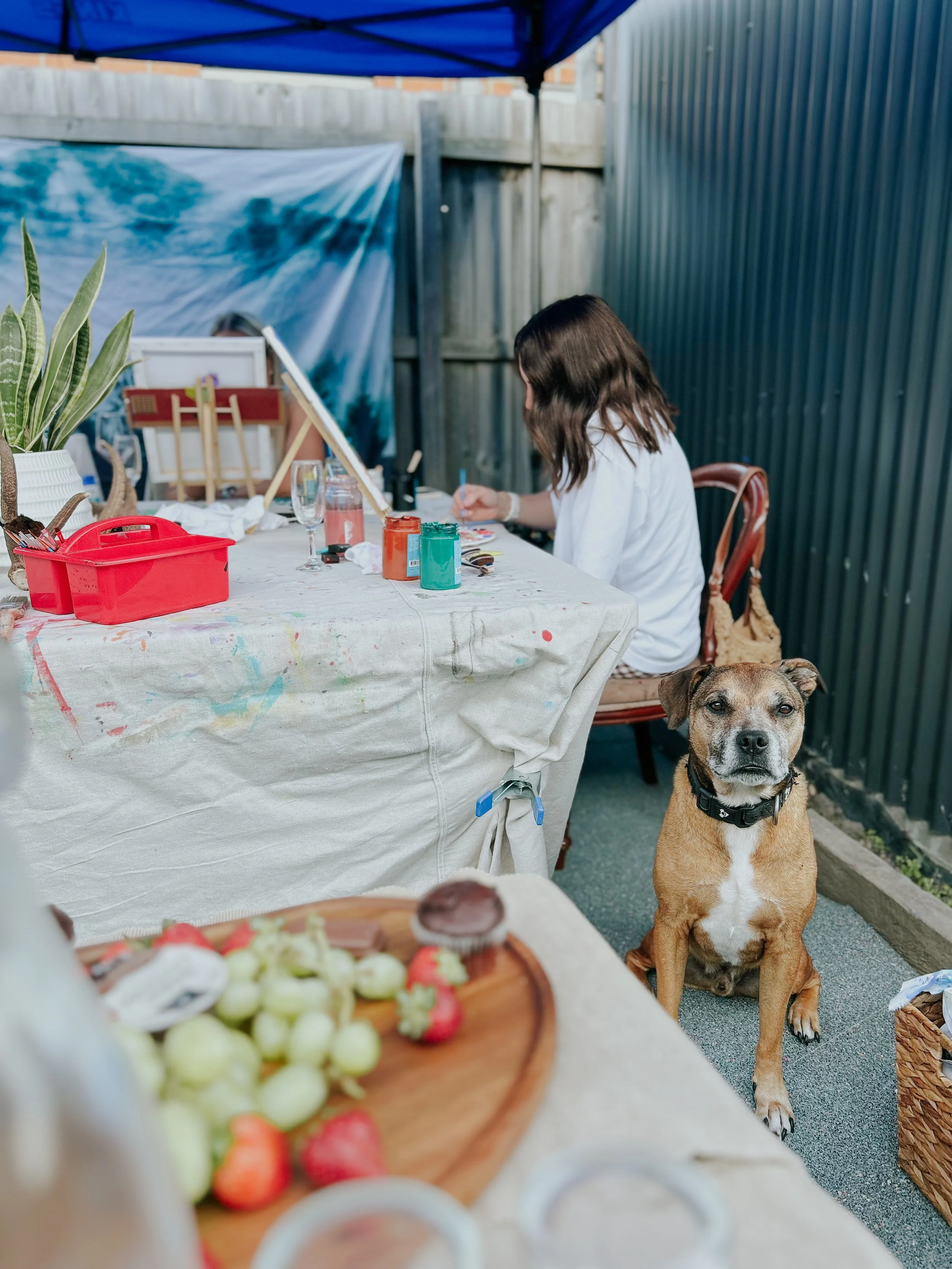 A woman painting at an outdoor art station, with a dog sitting nearby and a blurred fruit platter in the foreground.