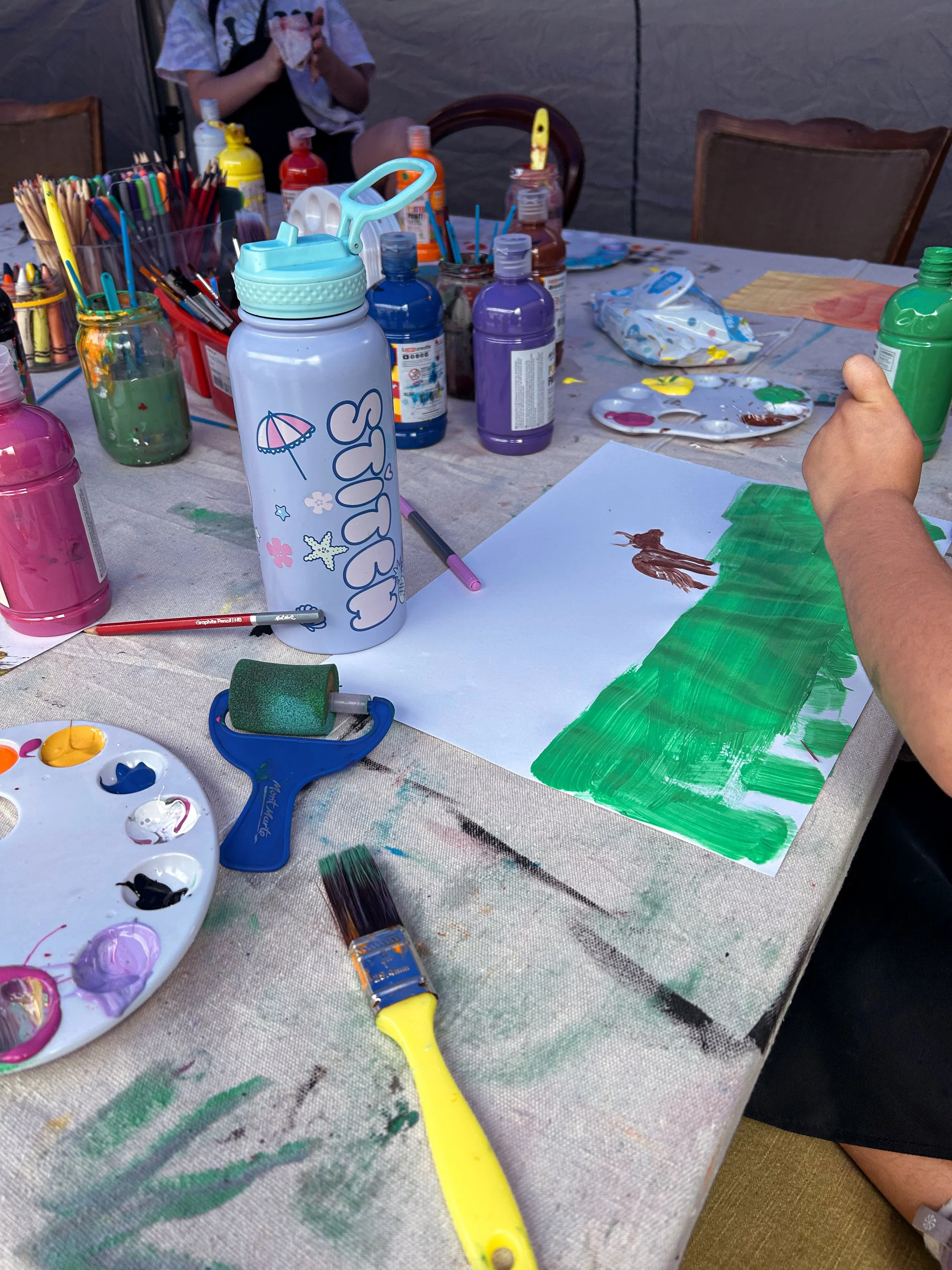 Child painting a picture of a person and trees with green paint on paper, surrounded by various bottles of paint, brushes, and art supplies on a table.