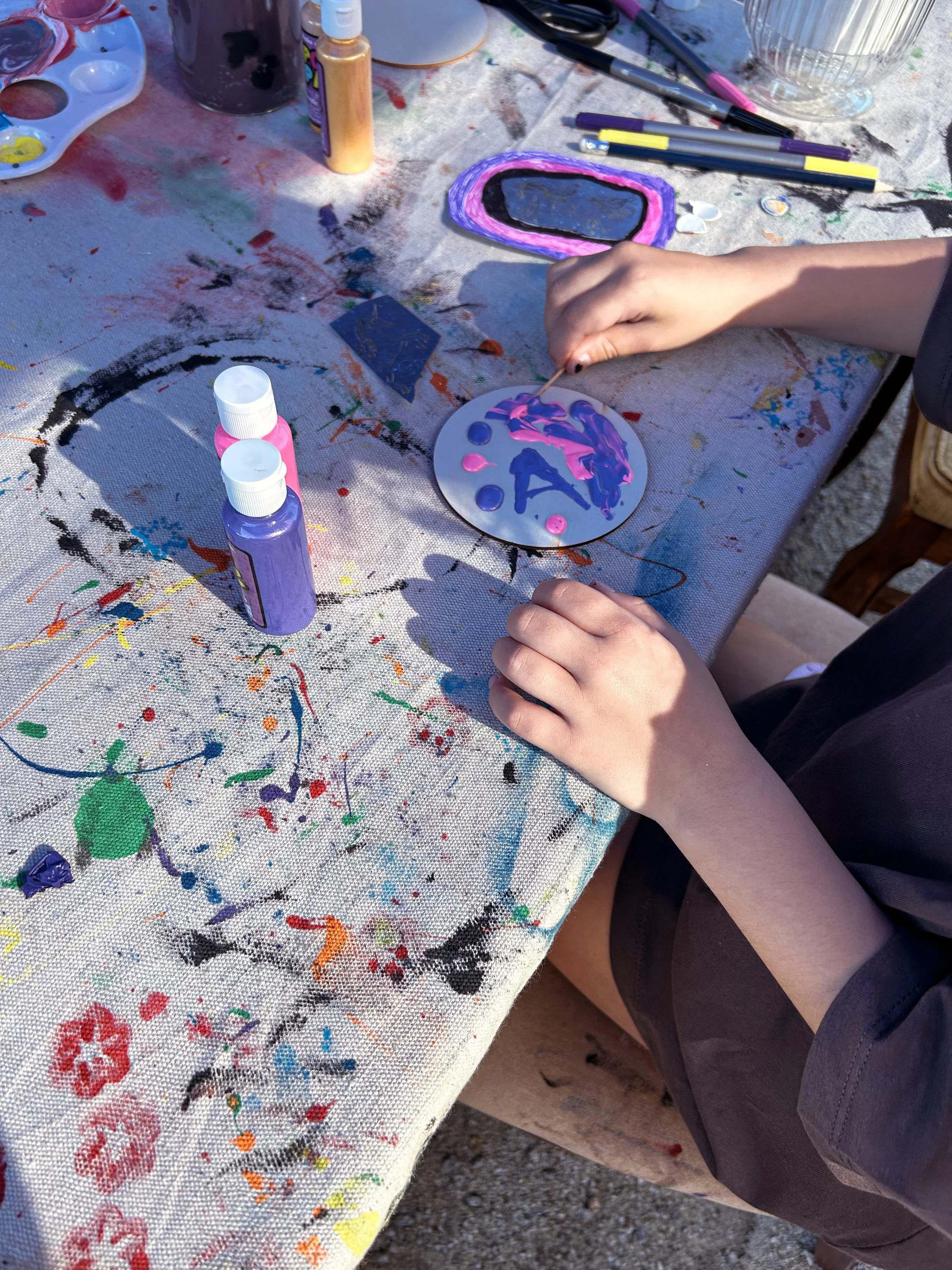 A person painting a decorative rock with purple, pink, and black paint. The table is covered with paint splatters and art supplies, including bottles of paint, brushes, and marking pens.