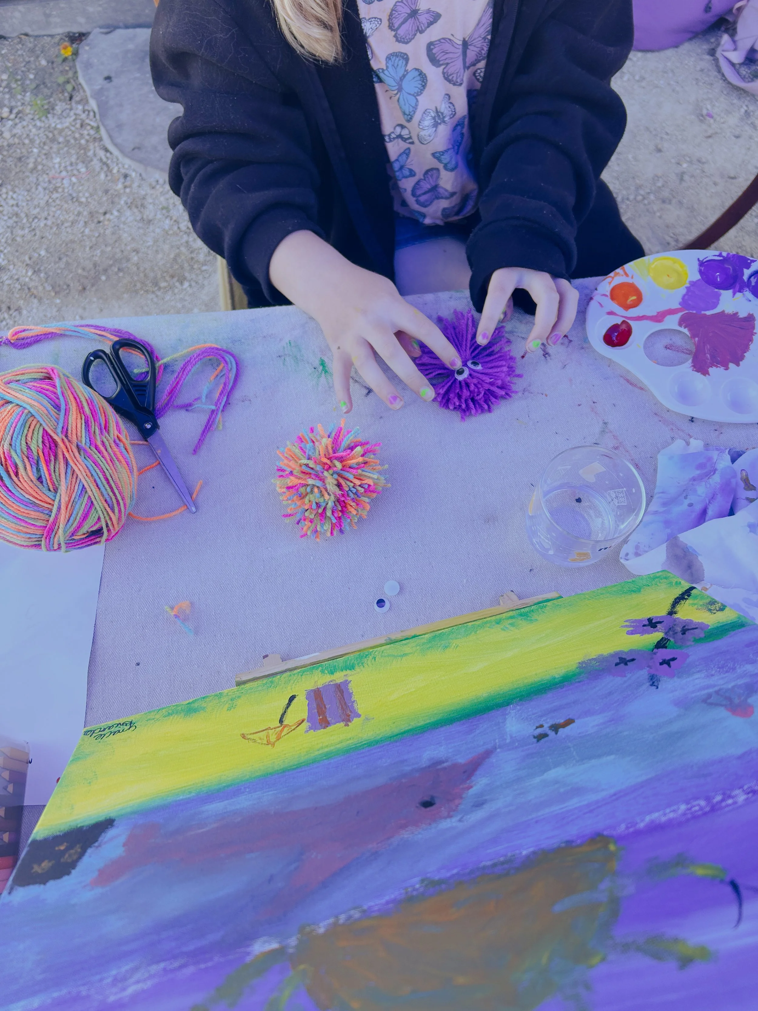 Person's hands creating colorful pom-pom craft at an outdoor arts and crafts table, with yarn, scissors, paint palette, and painted canvas visible.