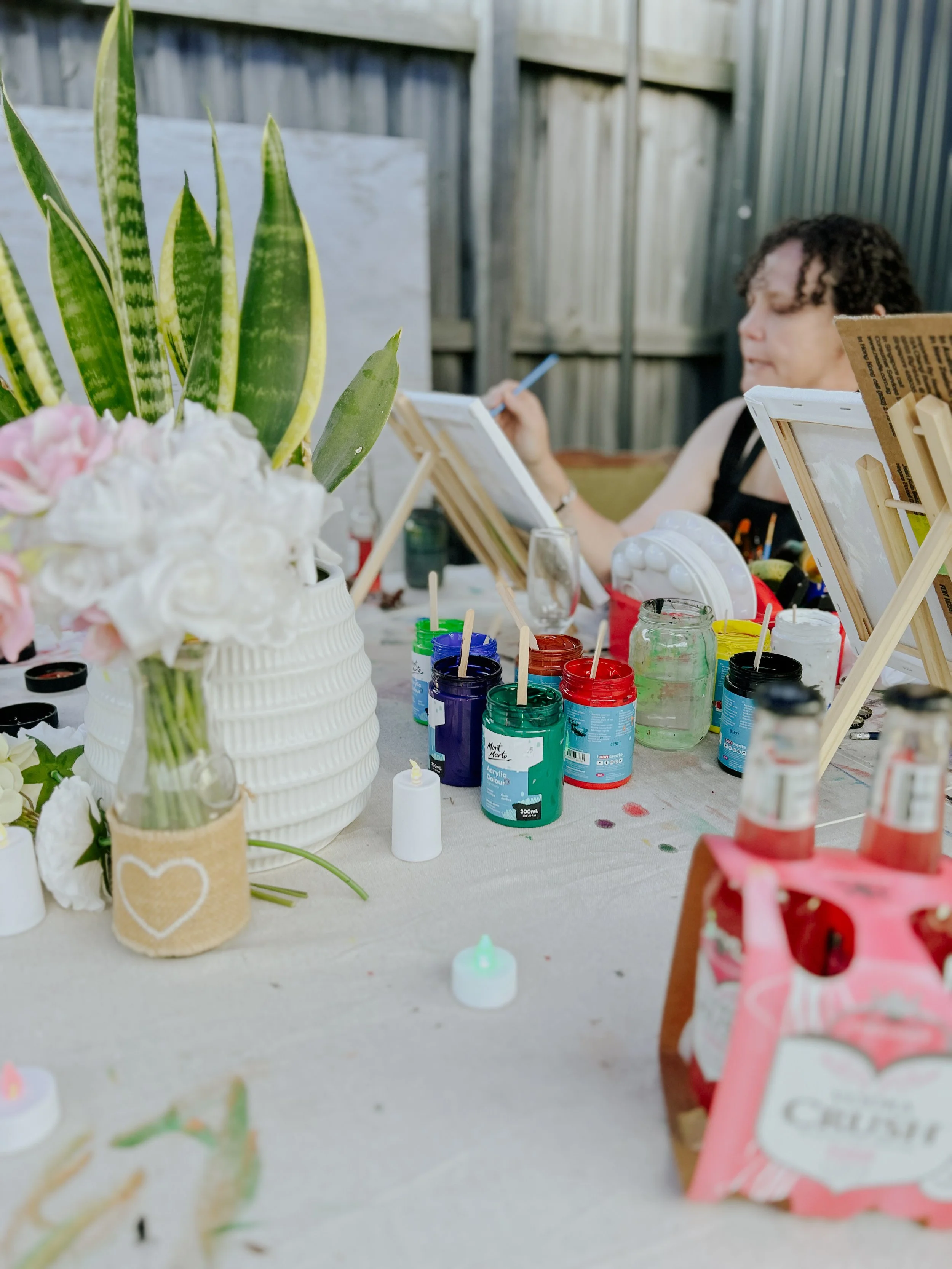 A woman sitting outdoors at a table with painting supplies, including open paint jars, brushes, and canvases, with large green plants and a wooden fence in the background.