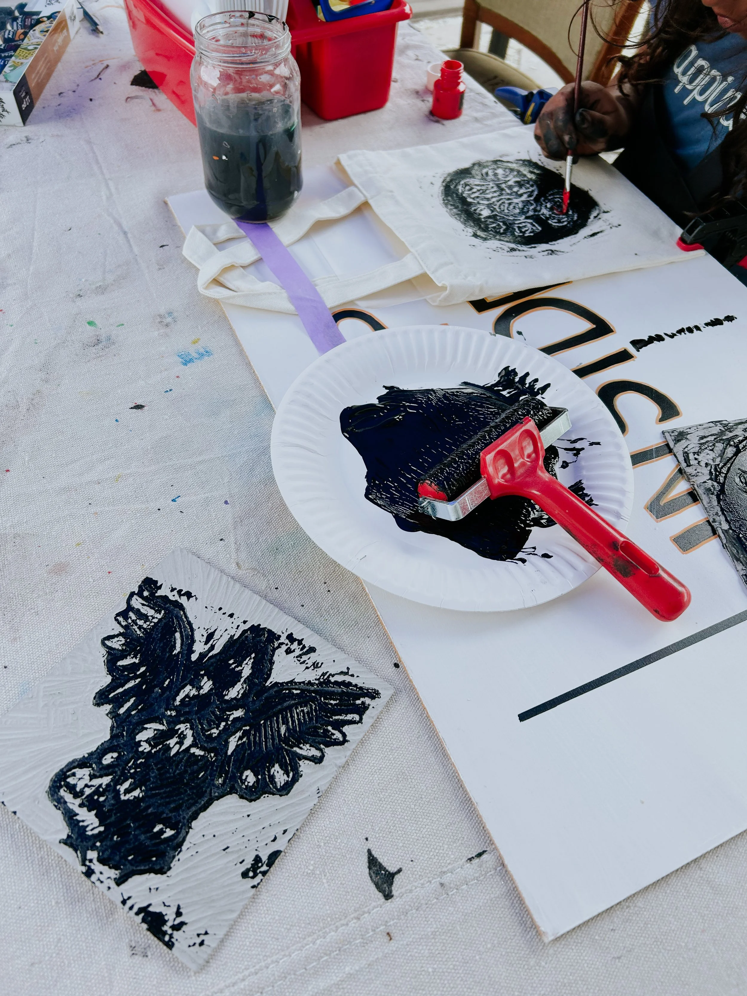 A craft activity setup with black ink, a brayer, artwork on paper napkins, a jar of water, and a workspace with paint and paper towels. Someone is using a brayer to make ink prints.