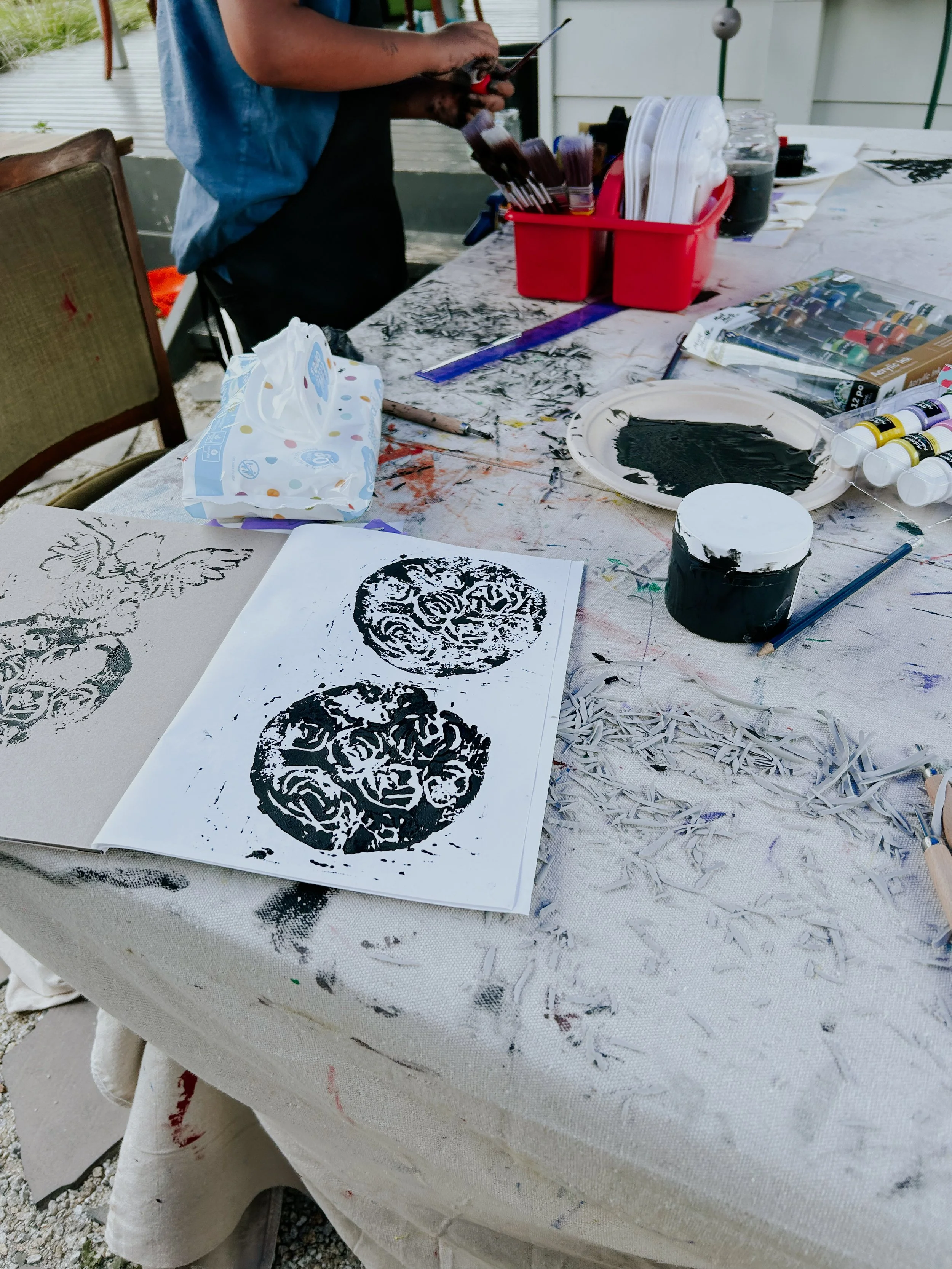 Arts and crafts workspace with paper prints of coffee cup designs, paint supplies, and a person in the background holding a tablet.