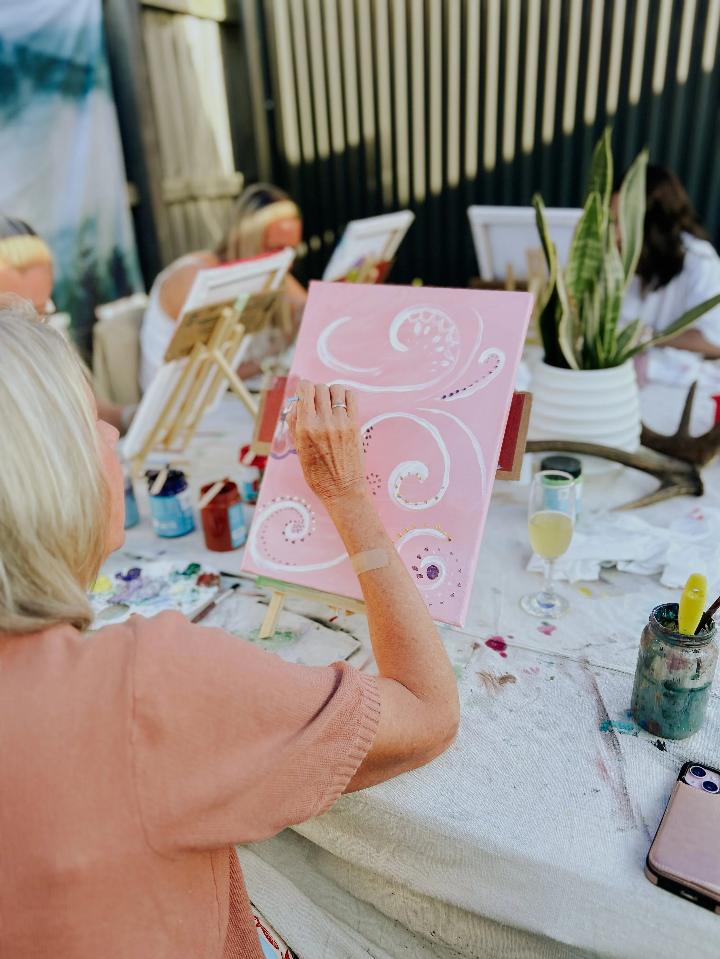 An elderly woman painting on a pink canvas with white and gold accents at an outdoor art event, surrounded by art supplies and other participants.