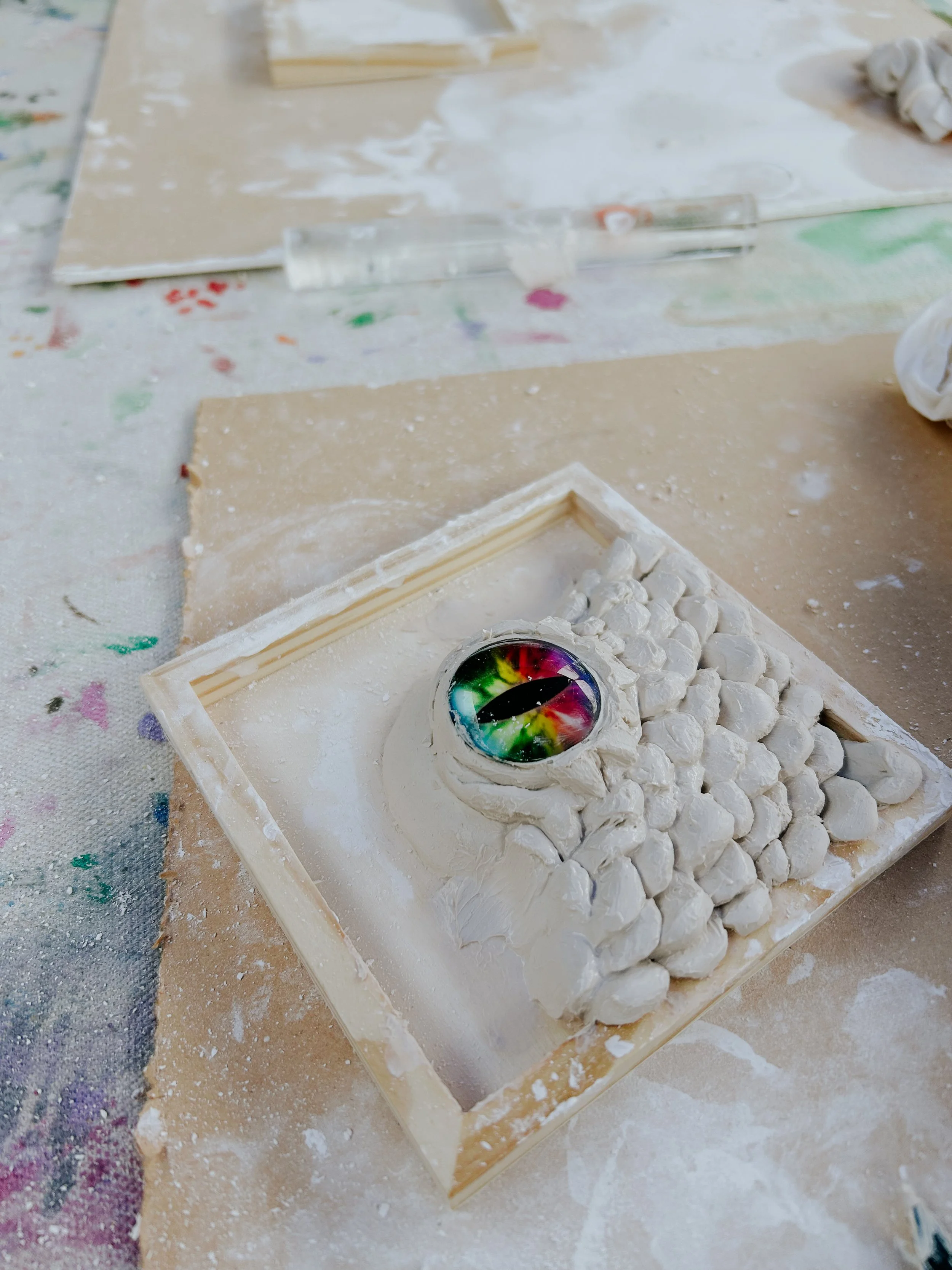 A partially completed ceramic art piece with a psychedelic rainbow-colored eye in the center, surrounded by white textured clay on a wooden frame, with a messy workspace including scattered paint splatters and tools.