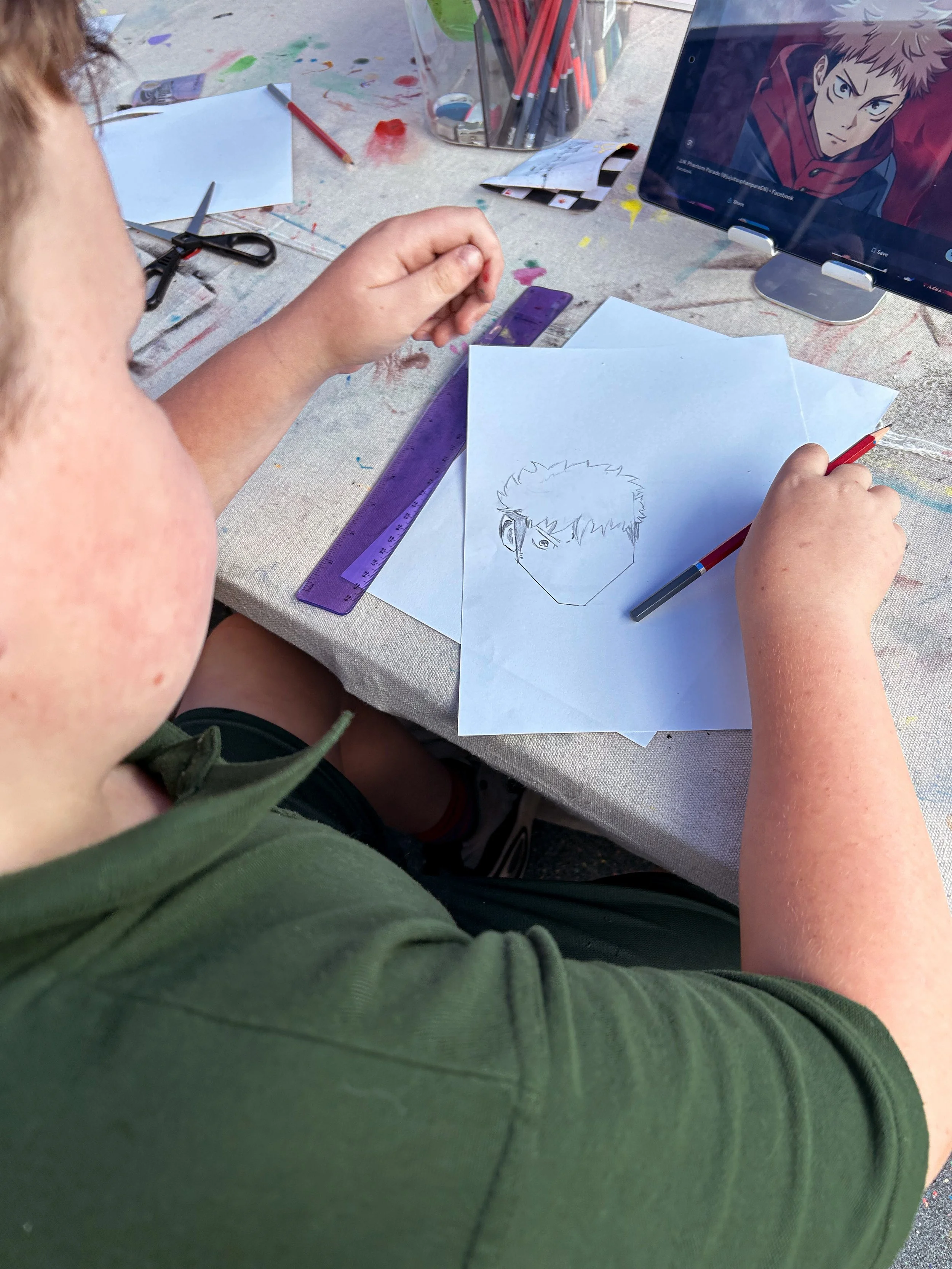 Child drawing a character's face on paper at an art table with colored pencils, scissors, and a tablet displaying a related character artwork nearby.