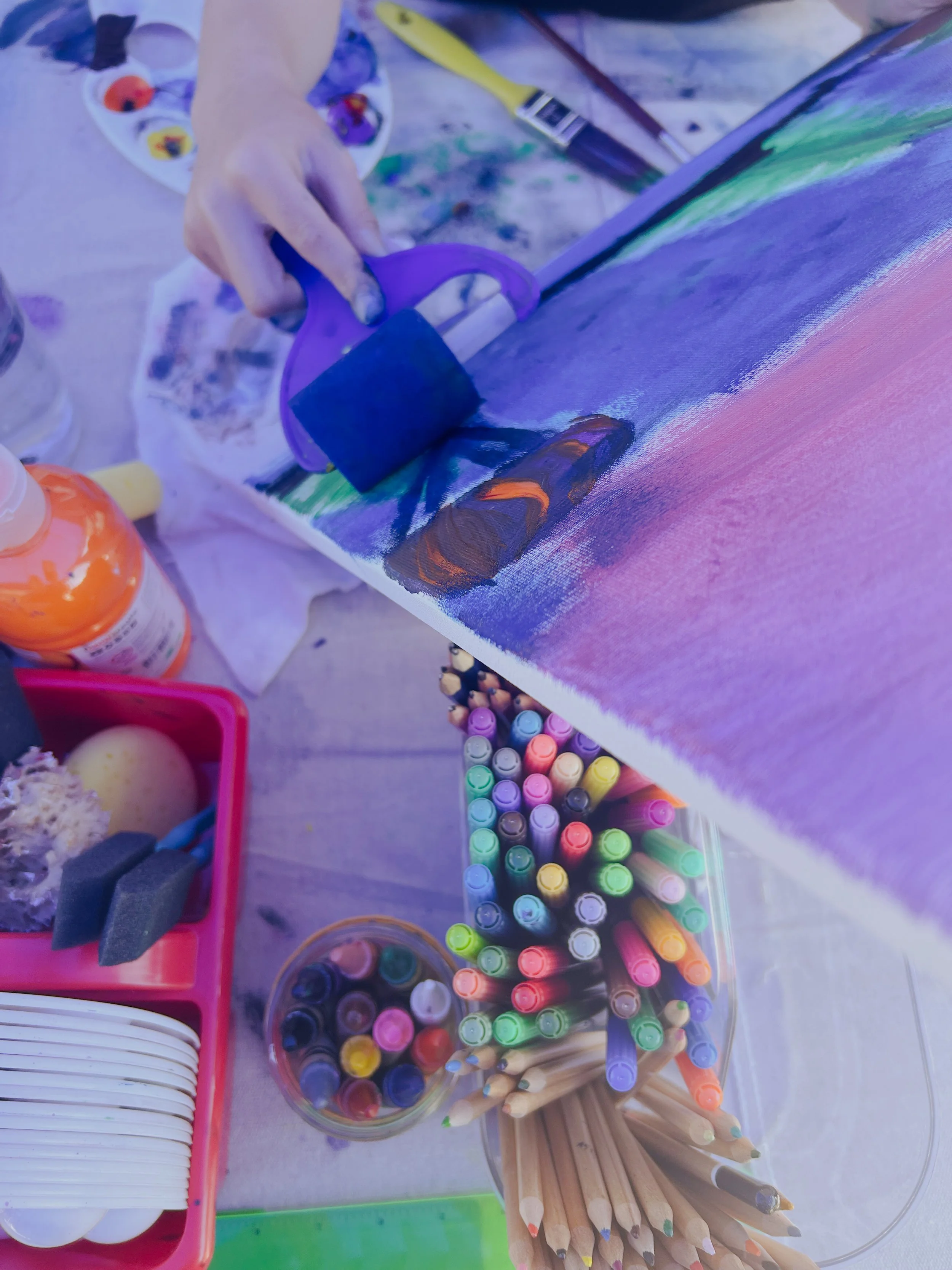 A person is painting a colorful landscape on a canvas using a roller brush. The table holds various art supplies including markers, colored pencils, paint bottles, and sponges, indicating an art activity.
