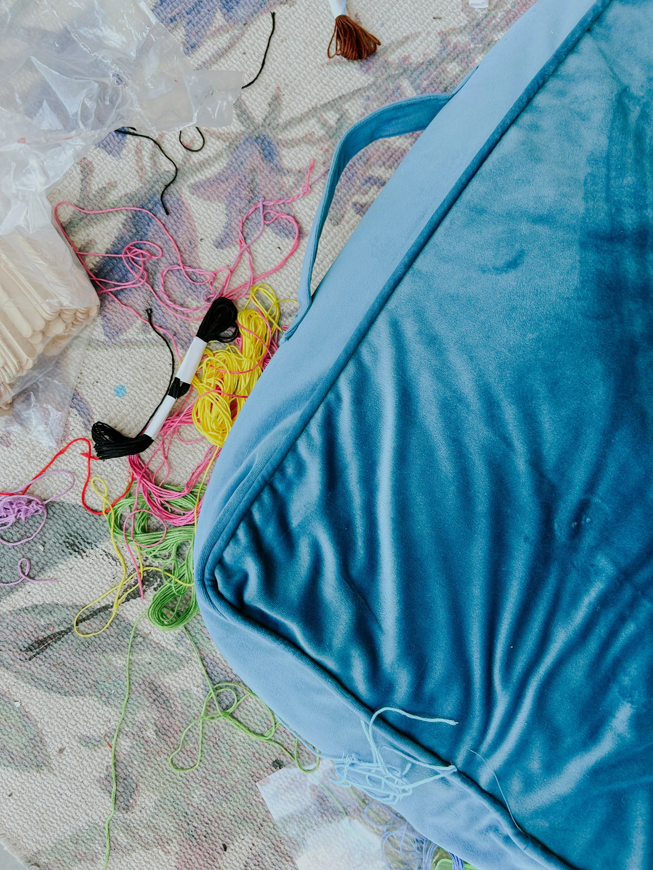 Multicolored yarn and knitting supplies scattered near a blue velvet furniture piece on a patterned rug.