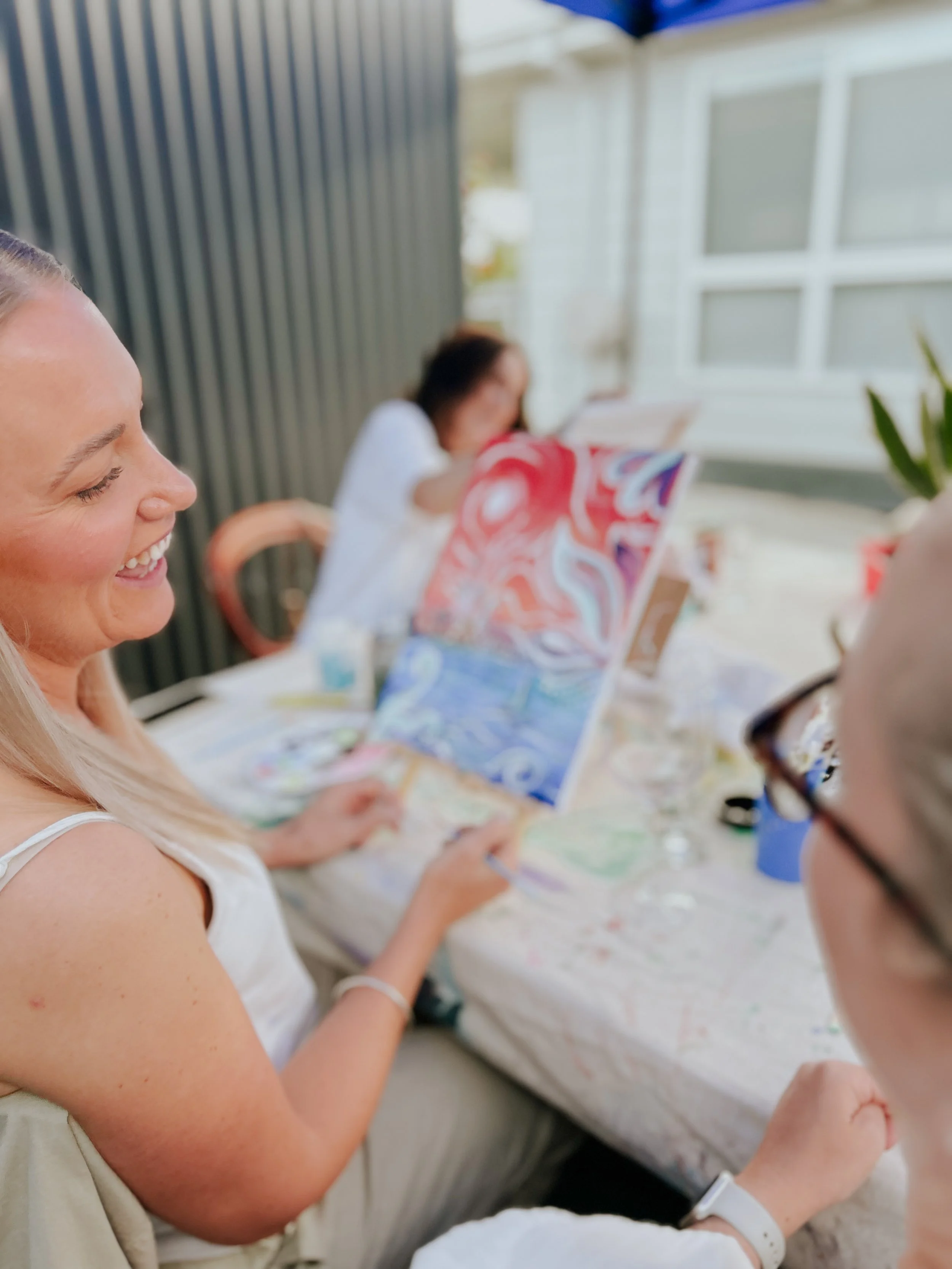 Women painting and enjoying art together outdoors, with colorful paintings on easels, on a table covered with paint supplies.