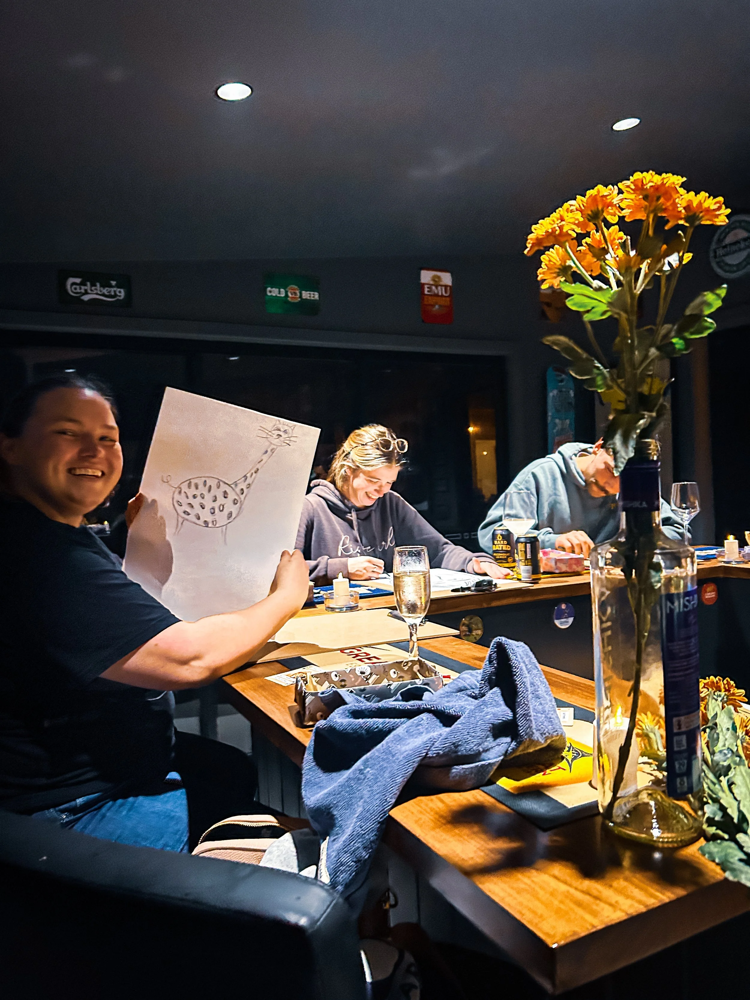 People sitting at a wooden table in a dimly lit room, with a person holding a drawing of a giraffe, a vase with yellow flowers, and various drinks.