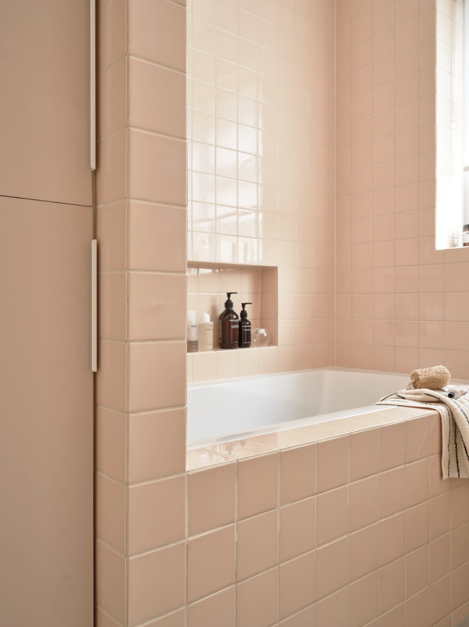 Bathroom with pink tiles, built-in shelf with soap and lotion bottles, white bathtub, beige towel, and a window with sunlight.