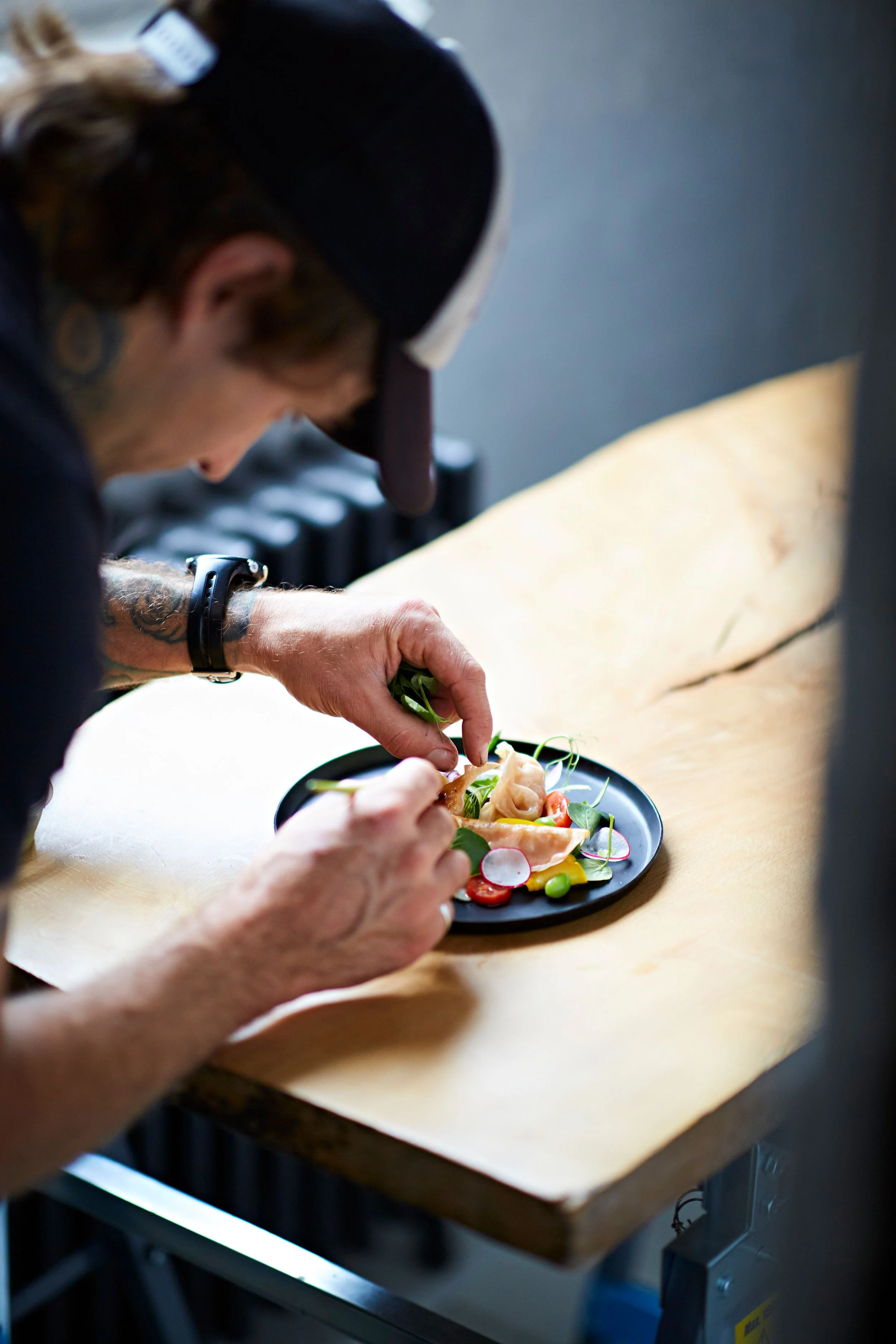 A chef or food preparer garnishing a dish with fresh herbs at a wooden work surface.