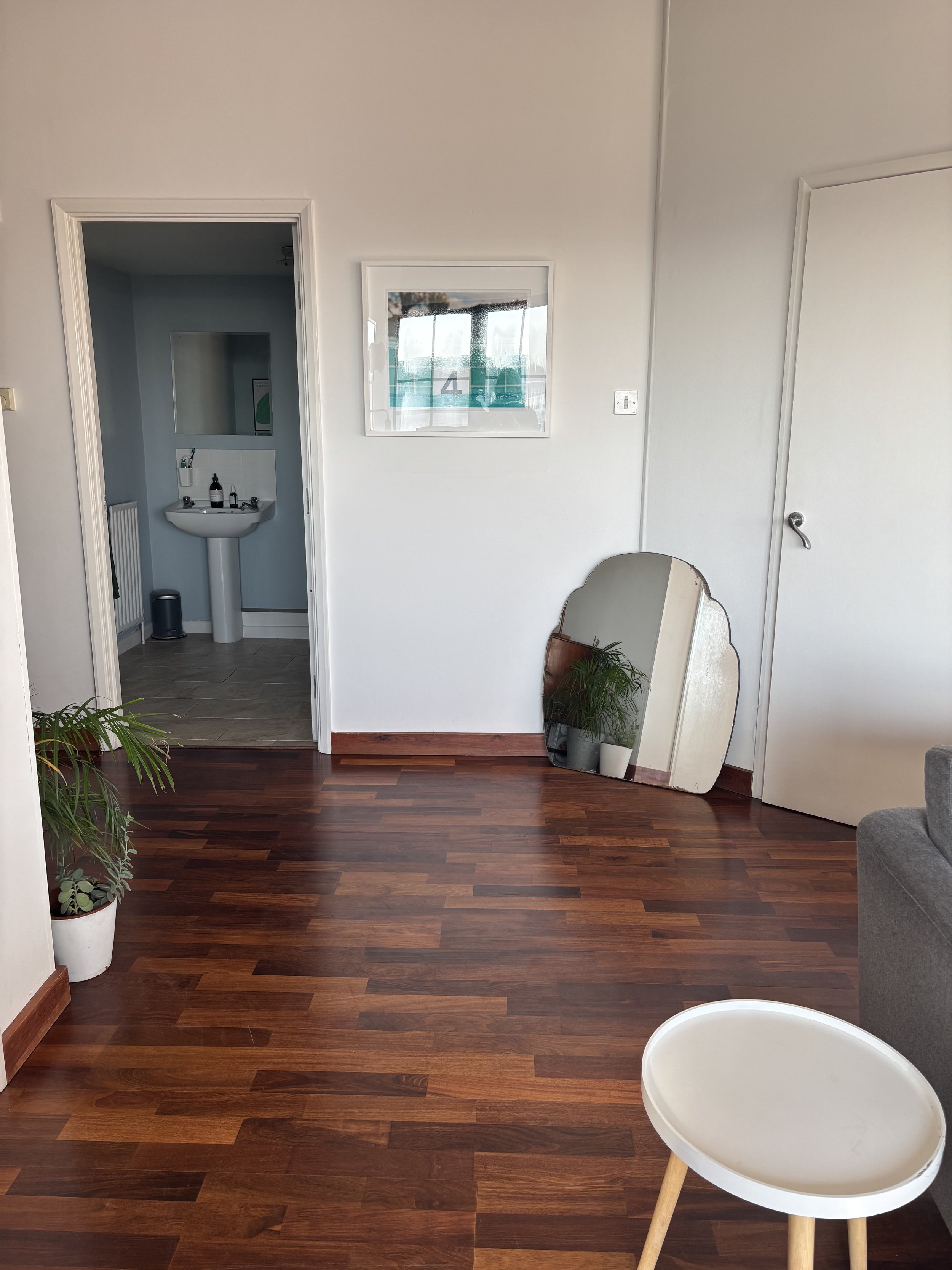 A living room with hardwood floors, two potted plants, a mirror leaning against the wall, and a small white table with wooden legs. Some plants and a mirror are reflected in the window on the wall.