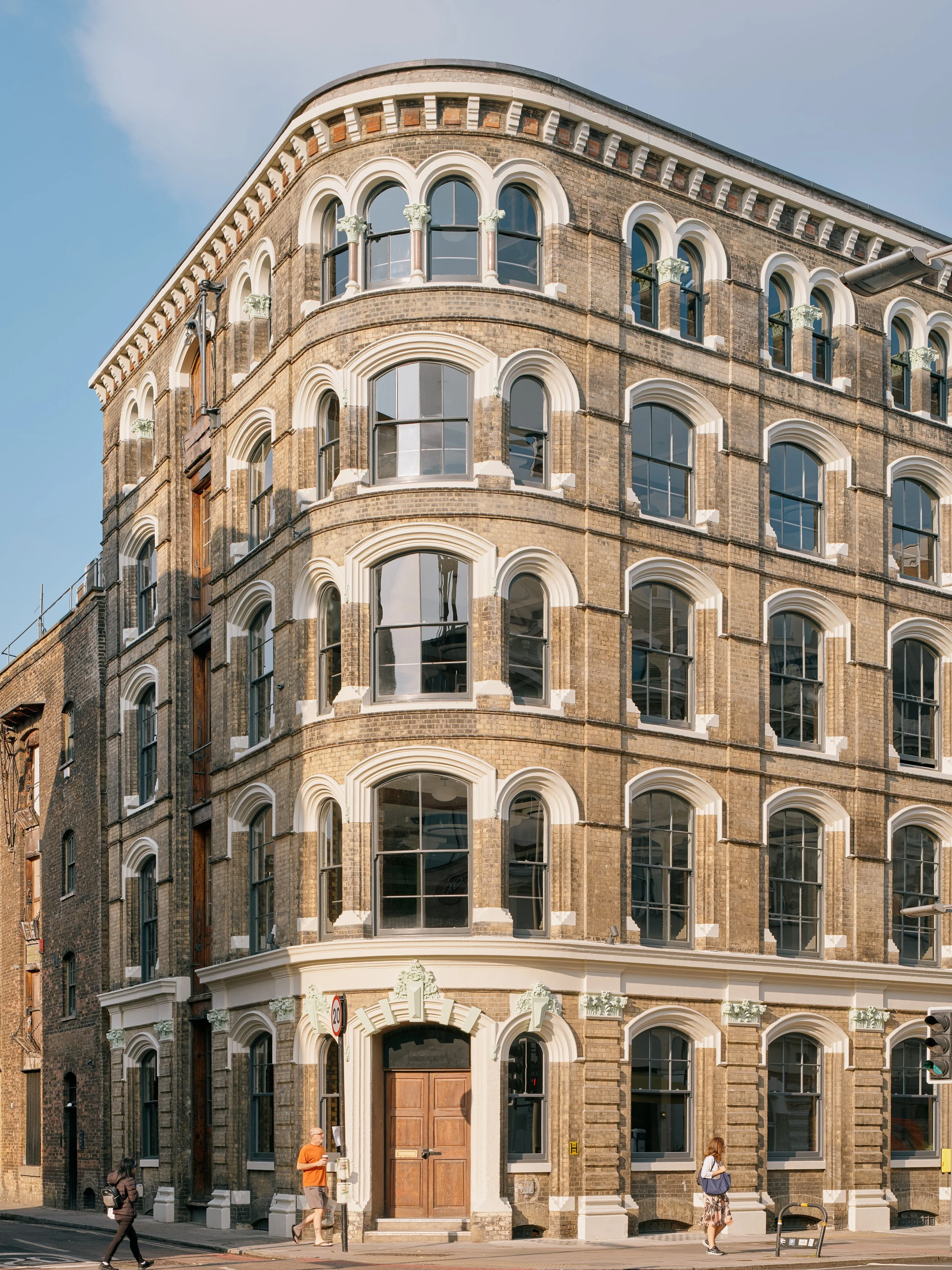 A multi-story brick building with arched windows and white decorative accents in an urban setting, with pedestrians walking on the sidewalk in front.
