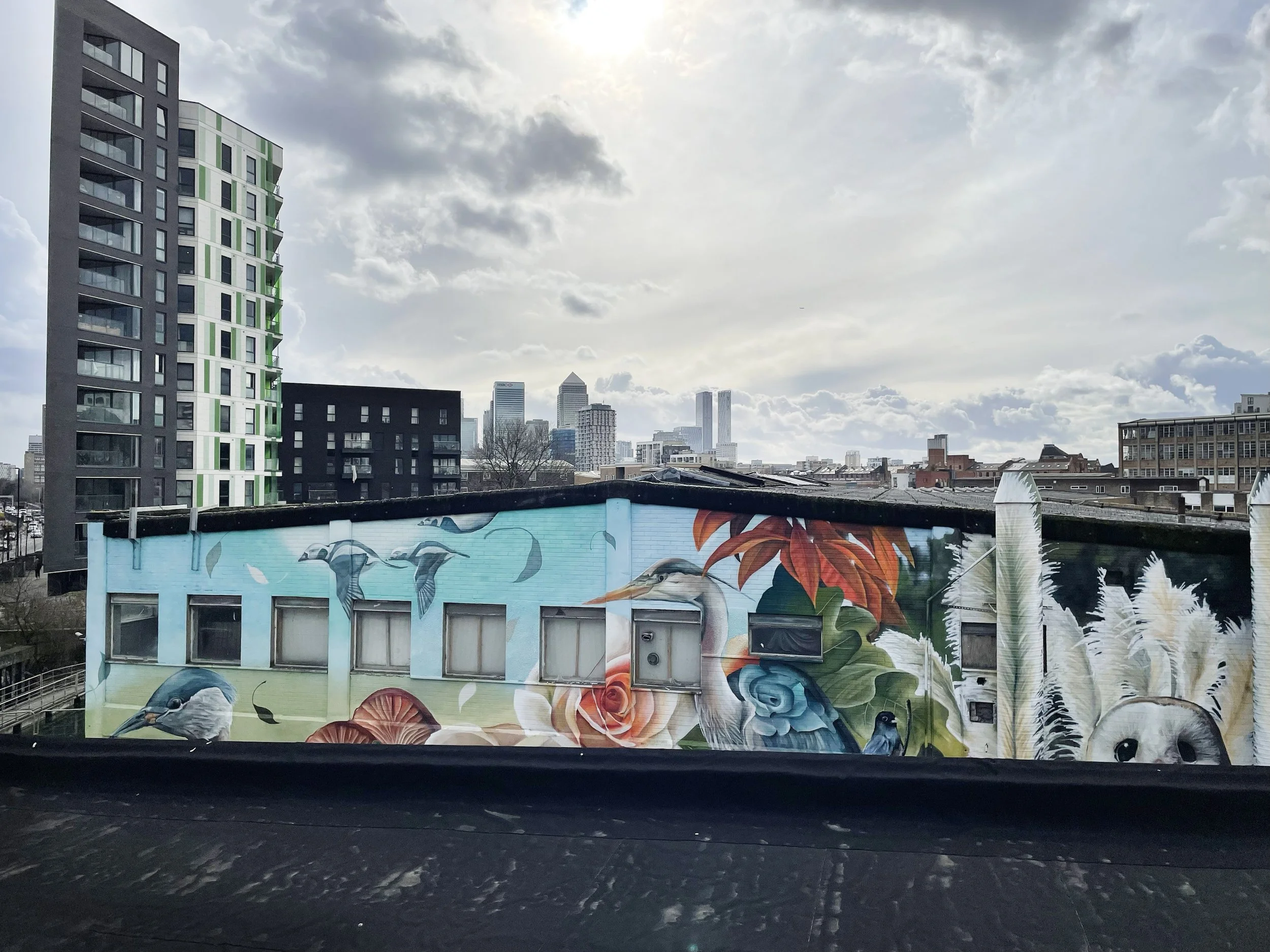 Cityscape with high-rise buildings and a mural on a lower building featuring birds, flowers, and a panda face.