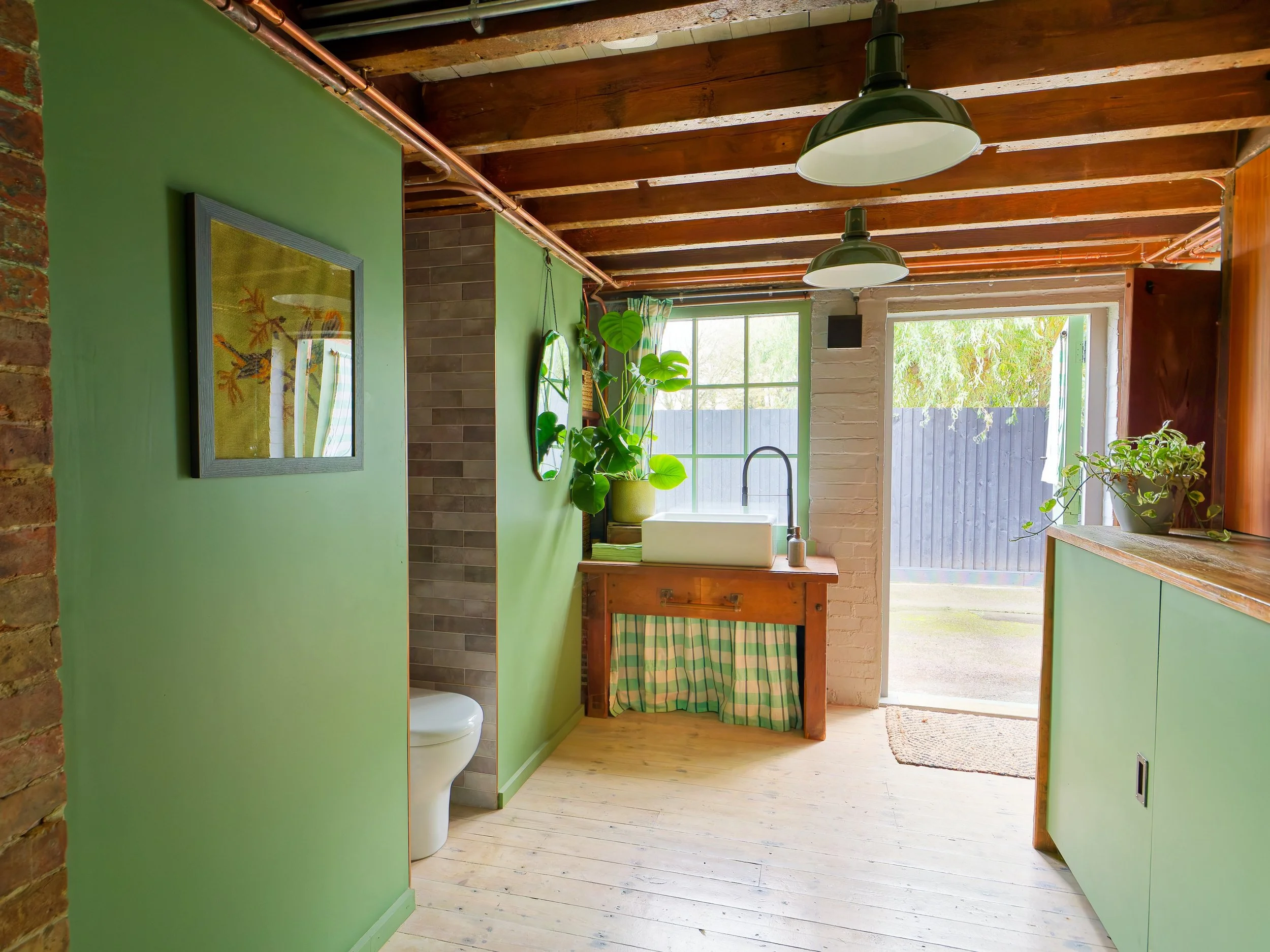A rustic-style room with green walls, wooden ceiling beams, and natural light from an open door. A small indoor plant sits on a counter with a white sink, and a soap dispenser. There are framed artworks and mirrors on the walls, and a toilet is parti