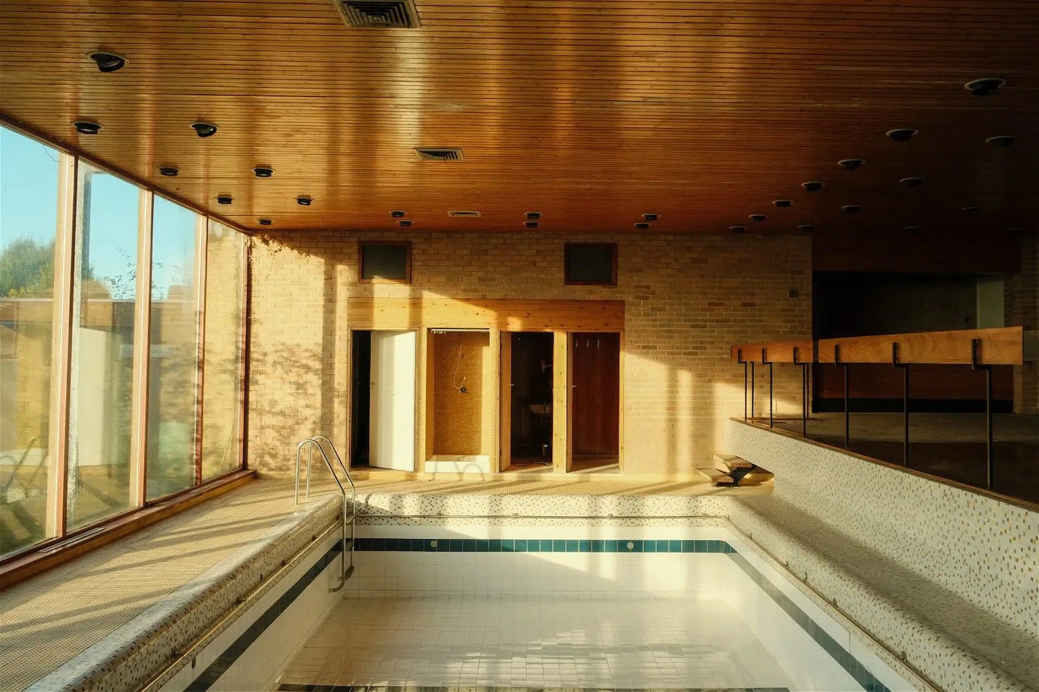 Empty indoor swimming pool with a ladder, large floor-to-ceiling windows, brick walls, and wooden ceiling, in a building with sunlight streaming in.
