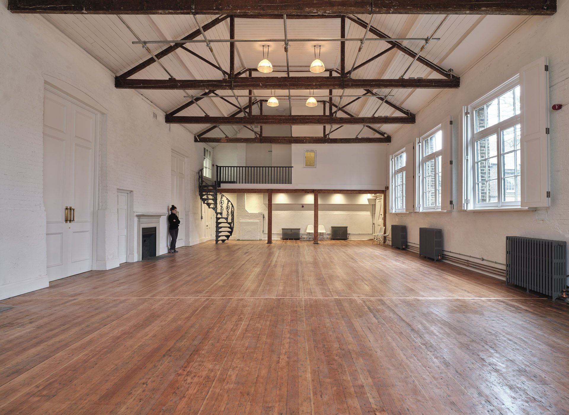 Spacious, empty room with wooden flooring, white brick walls, large windows with shutters, a high ceiling with exposed wooden beams, radiator heaters, and a small balcony or mezzanine accessible by a spiral staircase.