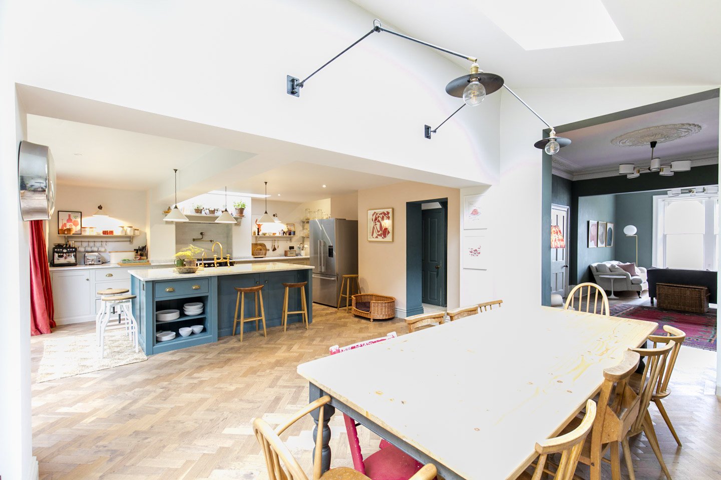 Open-concept kitchen and dining area with a large white table surrounded by chairs, blue kitchen island with stools, stainless steel refrigerator, and a living room with dark green walls, art, and sitting furniture.