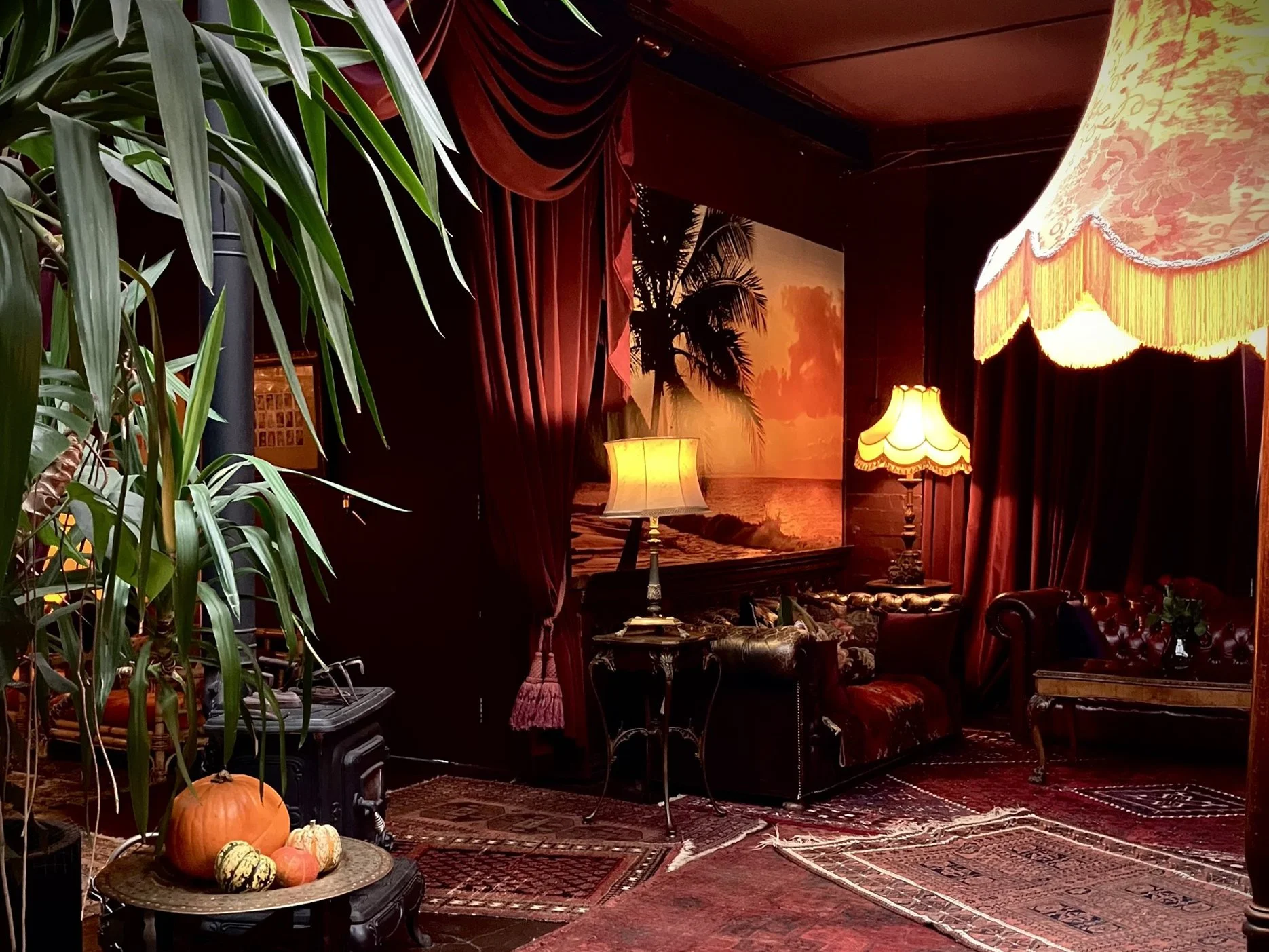 A cozy vintage living room with dark red curtains, antique lamps with fringed shades, a large tropical sunset painting, leather sofa, patterned rugs, and a pumpkin with gourds on a small table.