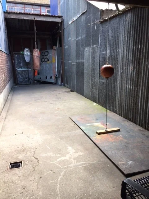 Side alley with a basketball mounted on a thin pole over a small wooden base, on a painted platform; corrugated metal fencing and an open garage with hanging punching bag and tools.