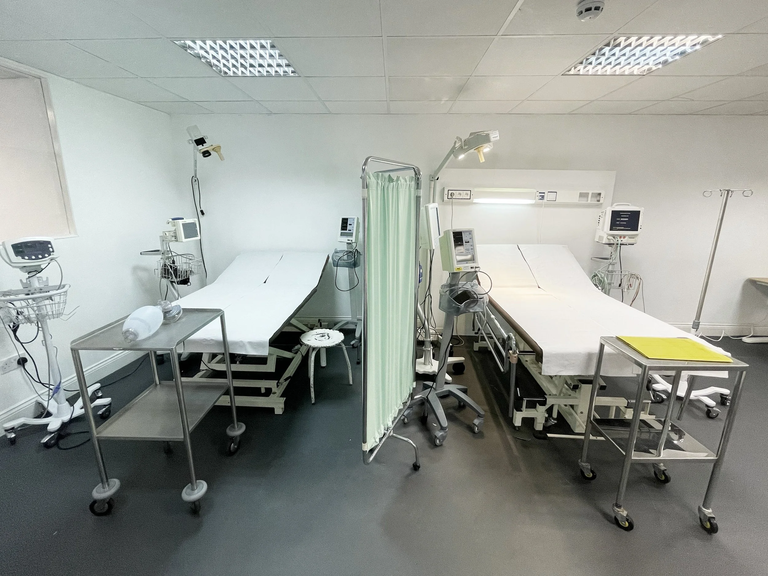 Empty hospital room with two beds separated by a green privacy curtain, surrounded by medical equipment, monitors, and mobile bedside tables.