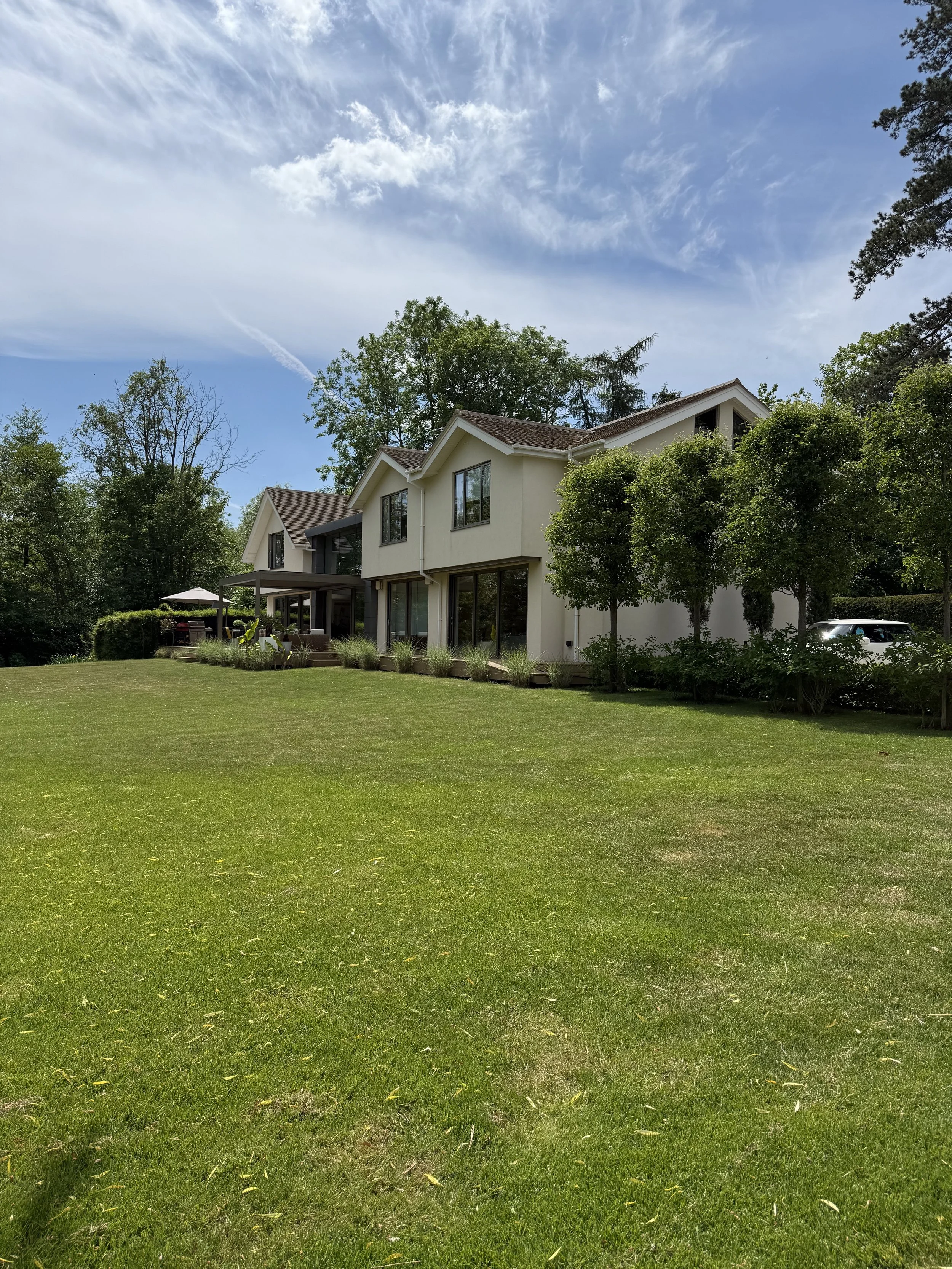 A modern white two-story house with large windows and a spacious lawn, surrounded by trees, under a partly cloudy blue sky.