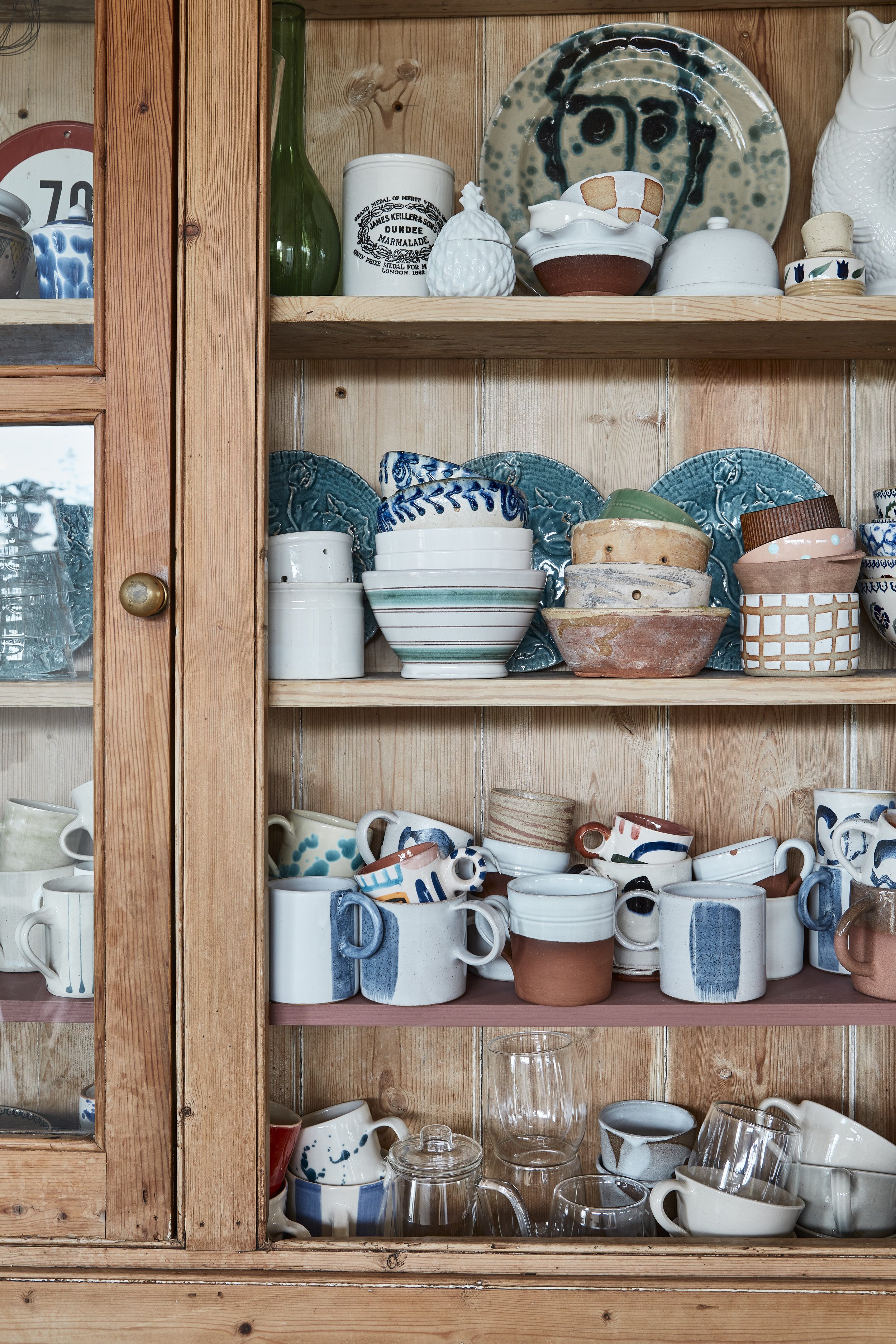 Wooden hutch with ceramic dishware, including plates, bowls, cups, and mugs in blue, white, and earthy tones, with some glassware and decorative items on the shelves.