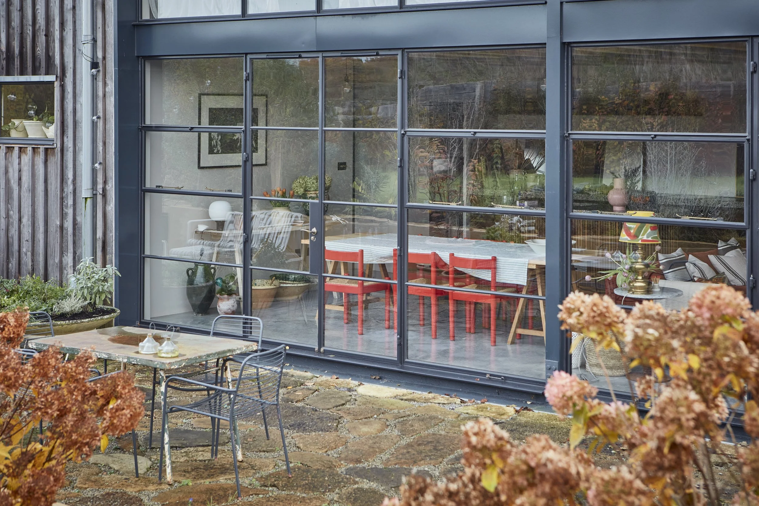 View of a modern house with large glass windows showing a decorated interior with a table and red chairs. Outside, there's a patio with a metal table and chairs, surrounded by plants with reddish-brown leaves.