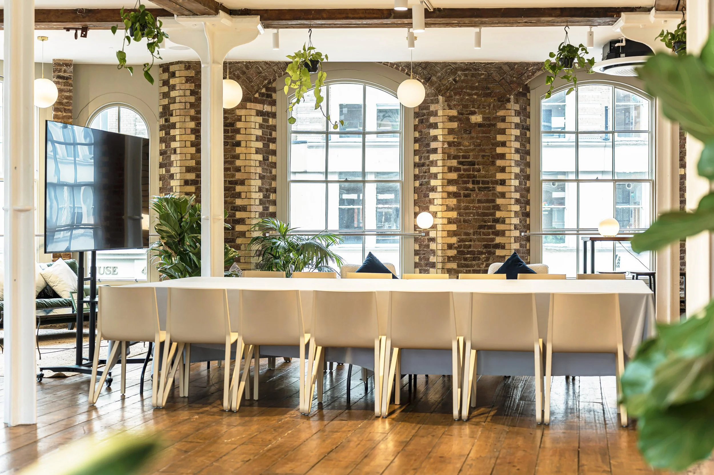 Modern interior space with brick walls, large windows, hanging plants, round pendant lights, a long table with white chairs, and indoor plants.