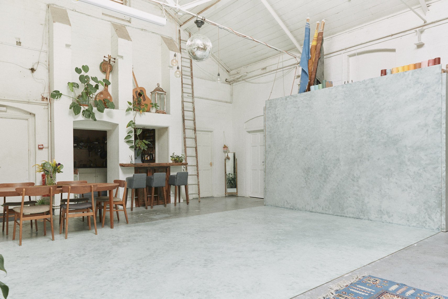 Indoor studio with white walls, a ladder, a disco ball, a bar area with barstools, a long dining table with chairs, plants, artwork, and a large textured partition.