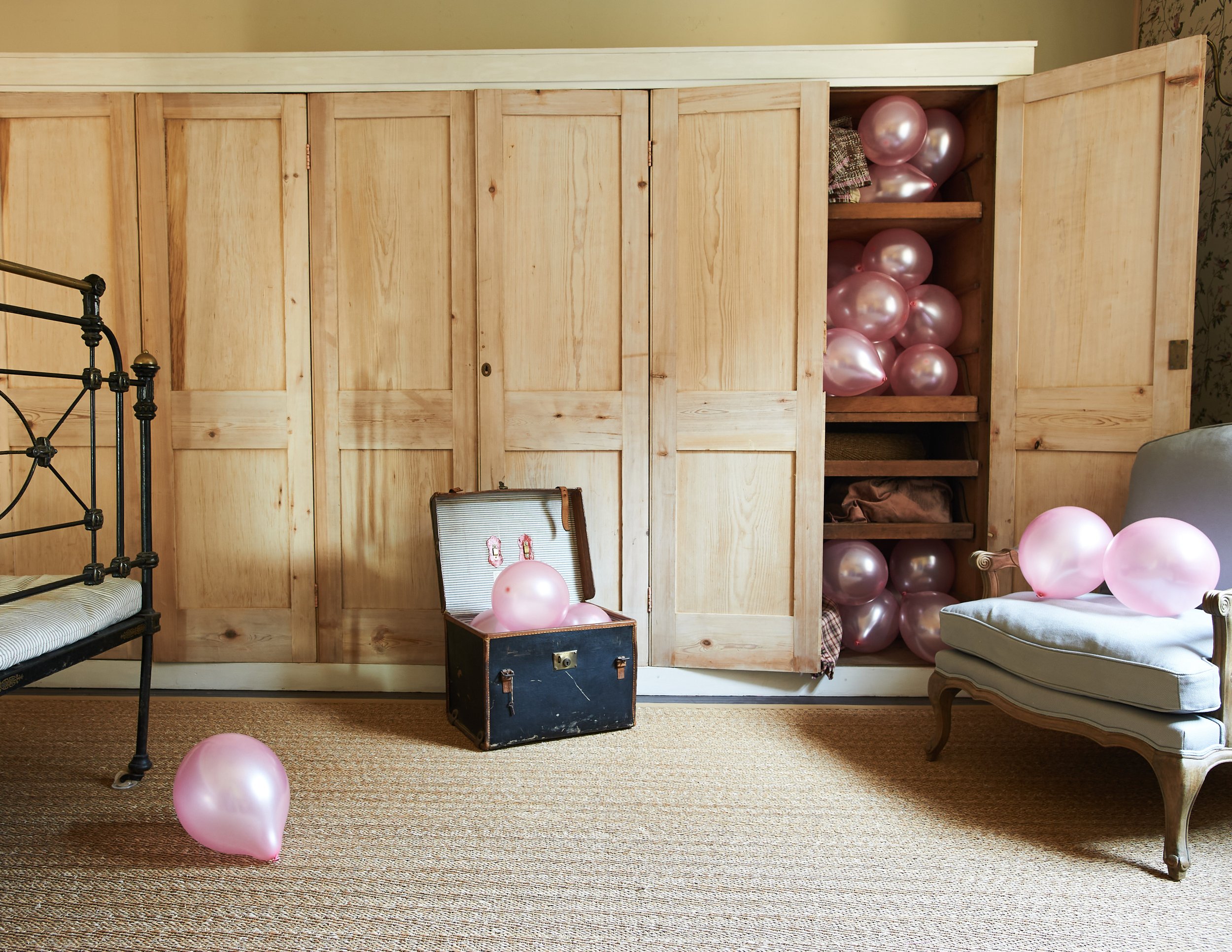 A vintage room with pink balloons, an open suitcase, and a wooden wardrobe filled with balloons and clothing.