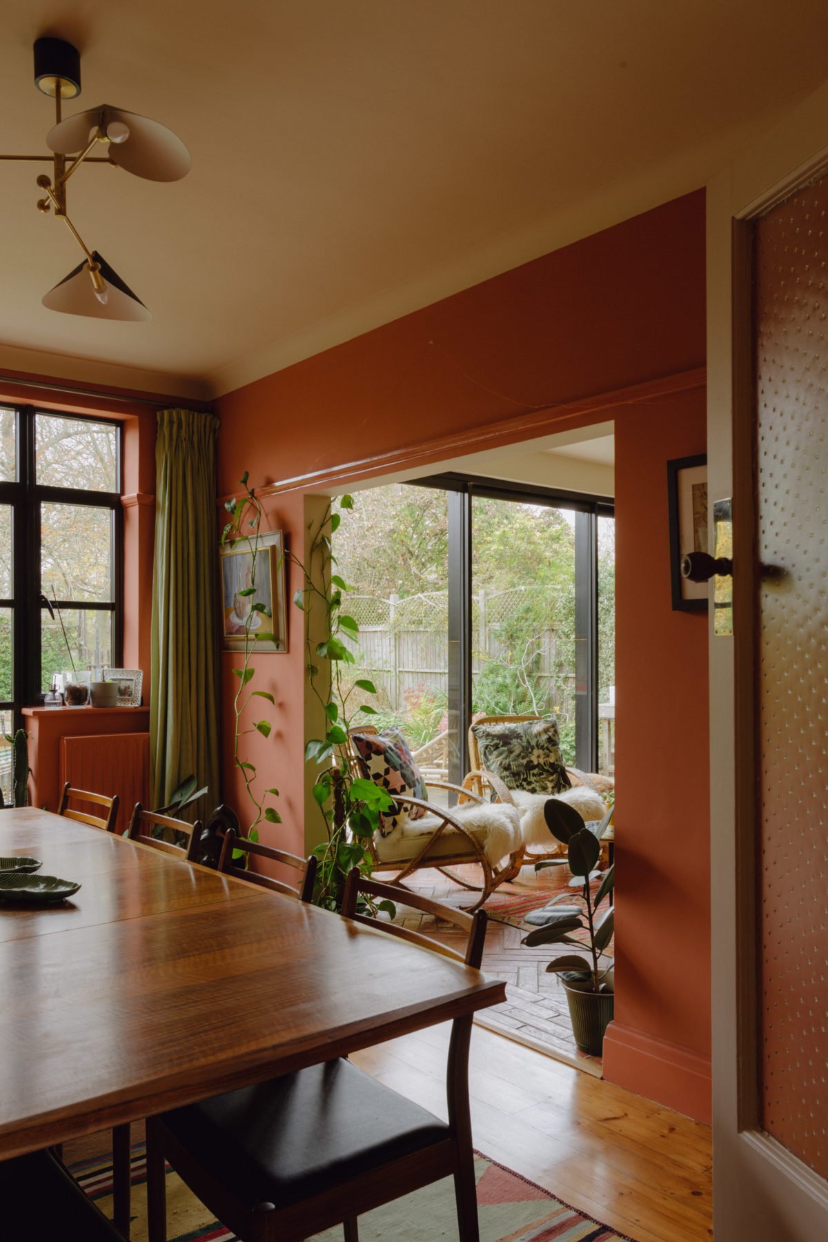 A cozy dining and sitting area with a wooden dining table, chairs, a large window, and a sunlit sitting nook with wicker chairs, potted plants, and outdoor views.