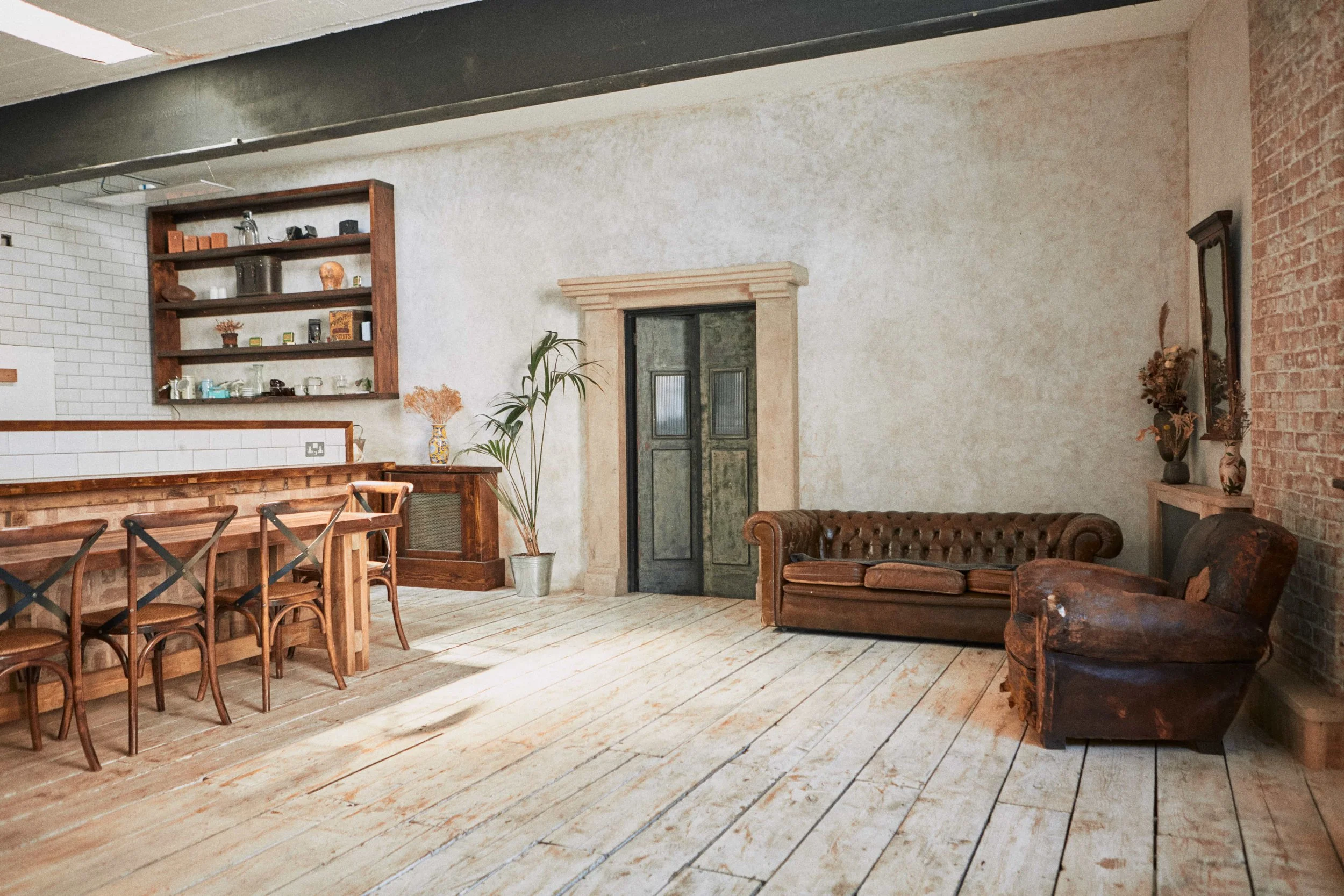 Living room with wooden flooring, brown leather sofas, a brick and white wall, a wooden dining table with chairs, a shelf with decorative items, and potted plants.