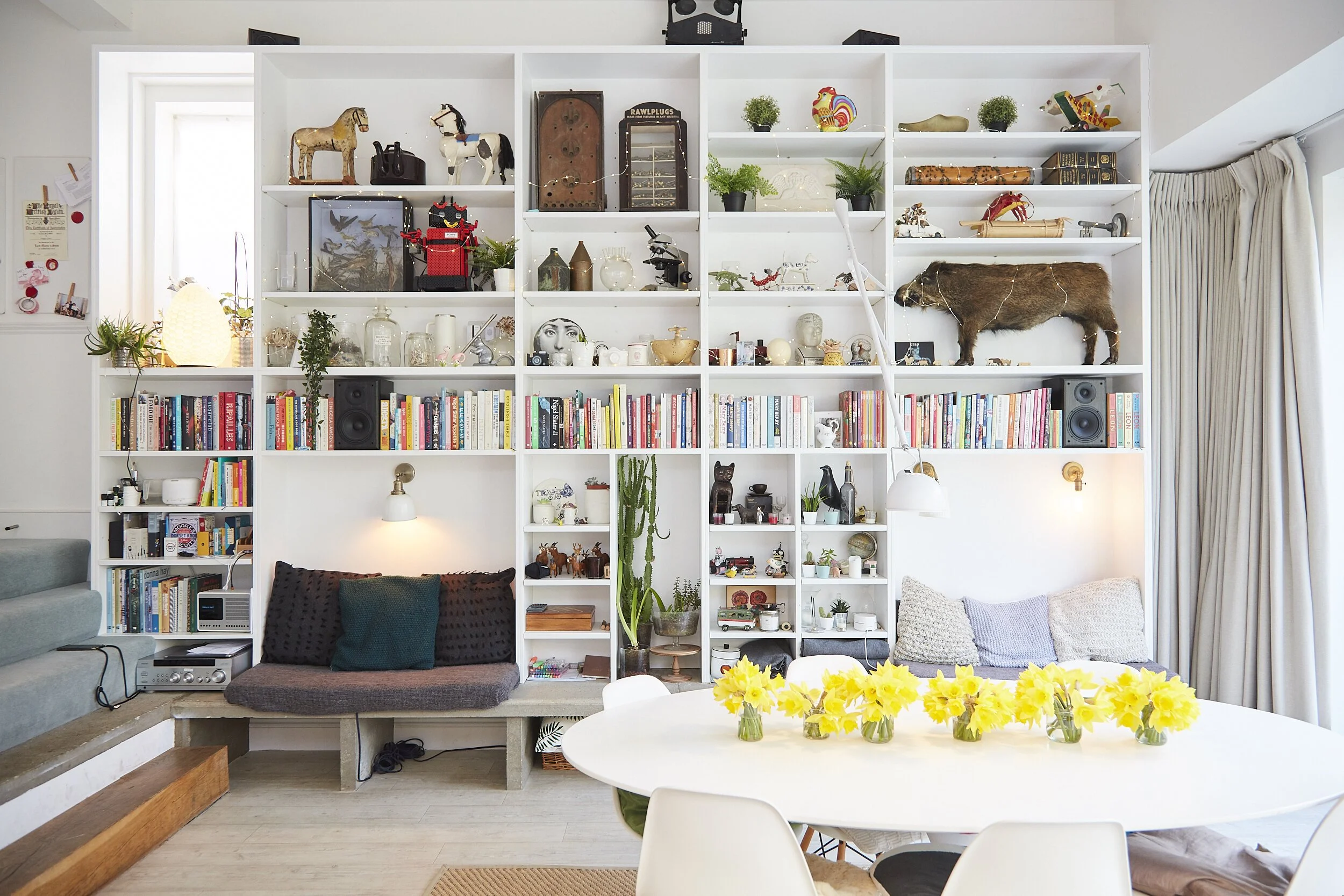 Living room with a large white bookshelf filled with books, decorative items, and plants, adjacent to a window with curtains. In front, a round table with yellow flowers and white chairs. There are couches and pillows, with a mix of decor including a