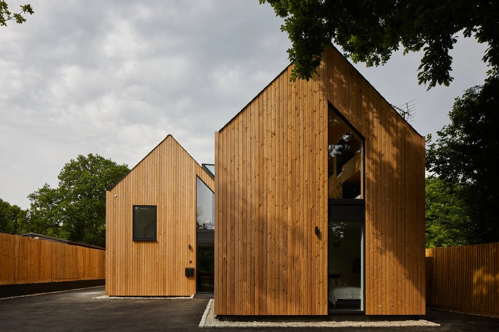 Modern wooden house with vertical slat siding, large glass windows, and a black door, surrounded by trees and a black driveway.