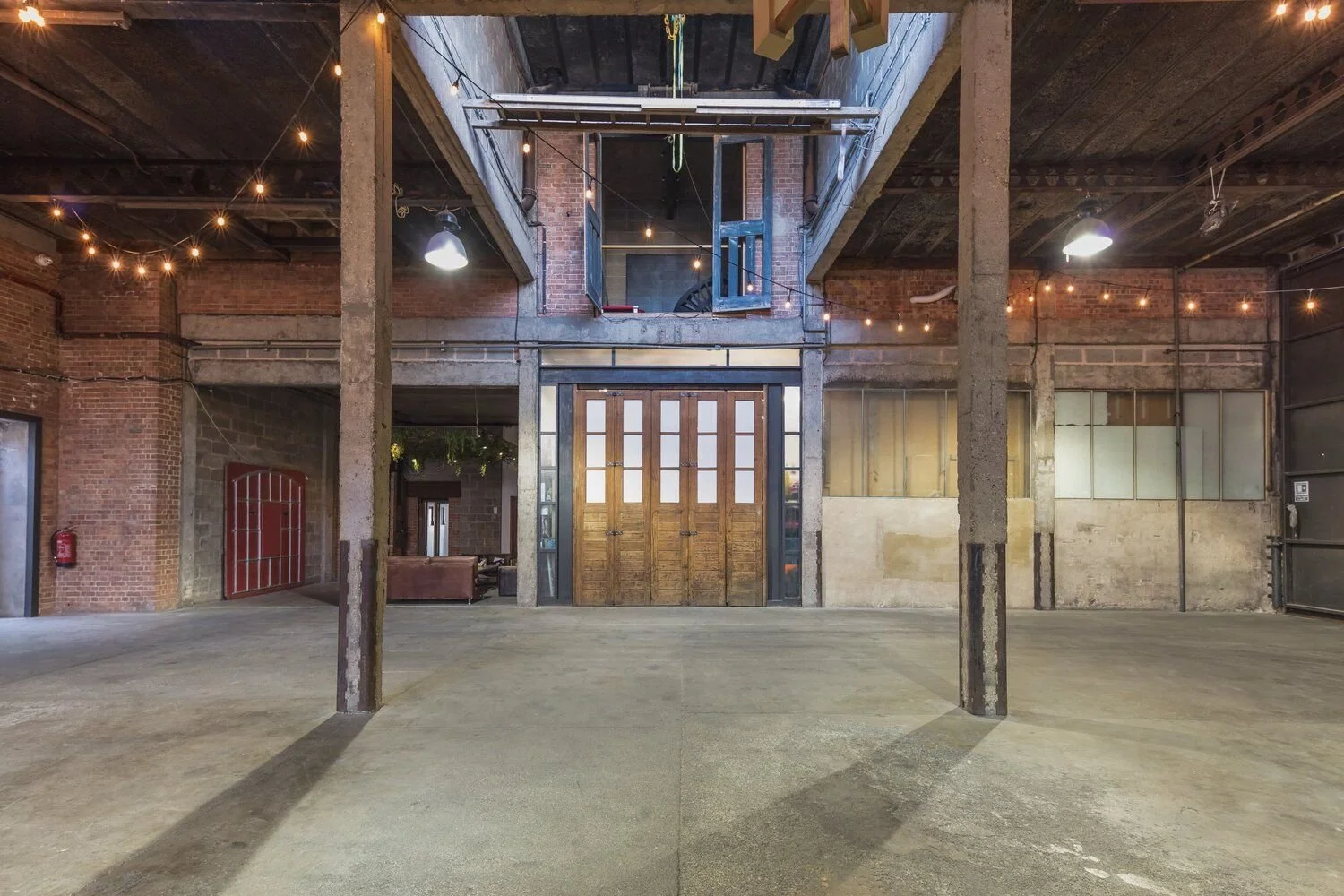 An industrial-style open space with exposed brick walls, concrete pillars, and a high ceiling. There are string lights hanging from above, a large wooden door at the center with glass panels, and a loft area above it with an open blue door. In the ba