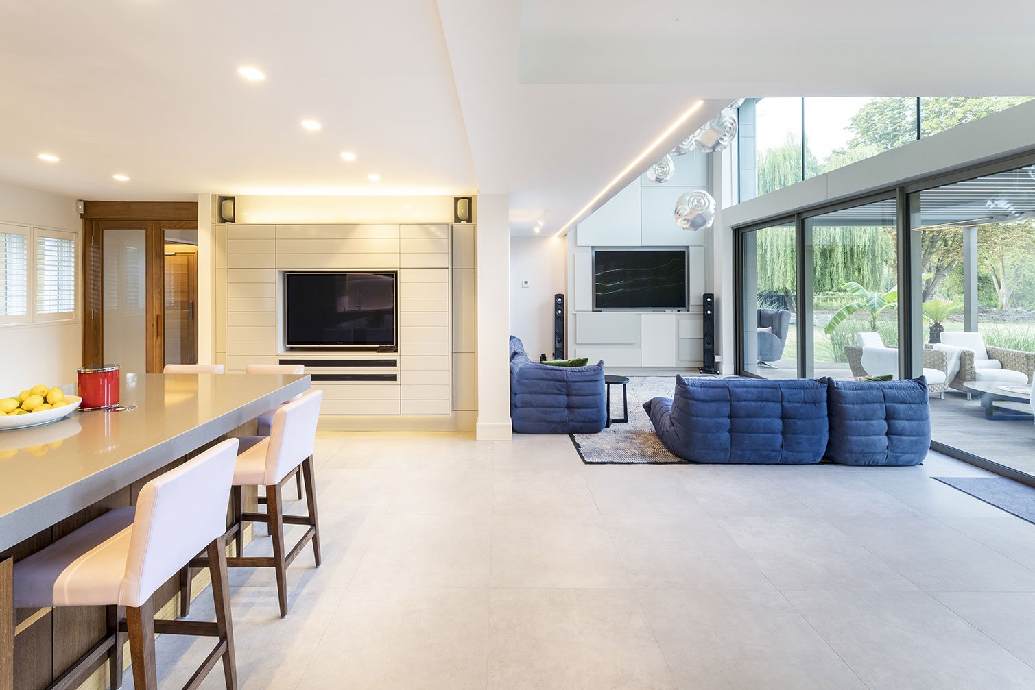 Modern living room with large glass sliding doors opening to outdoor patio, featuring blue sofas, flat-screen TVs, speaker systems, and minimalist decor.
