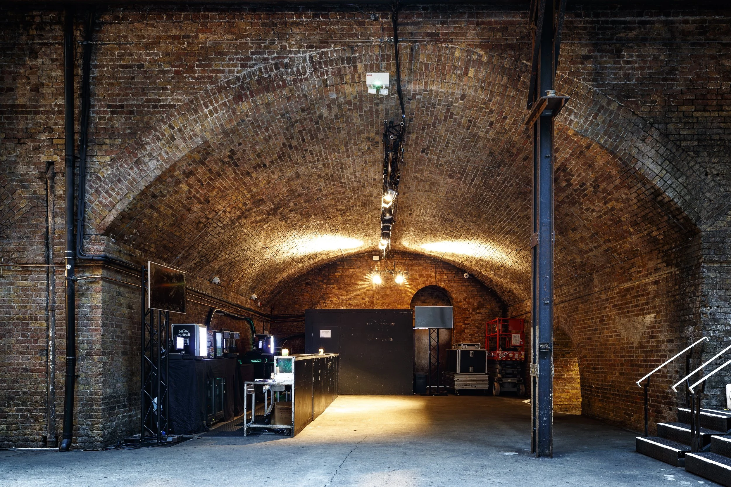 Interior of a brick tunnel or arch with lighting, equipment, and stairs, possibly a venue or industrial space.
