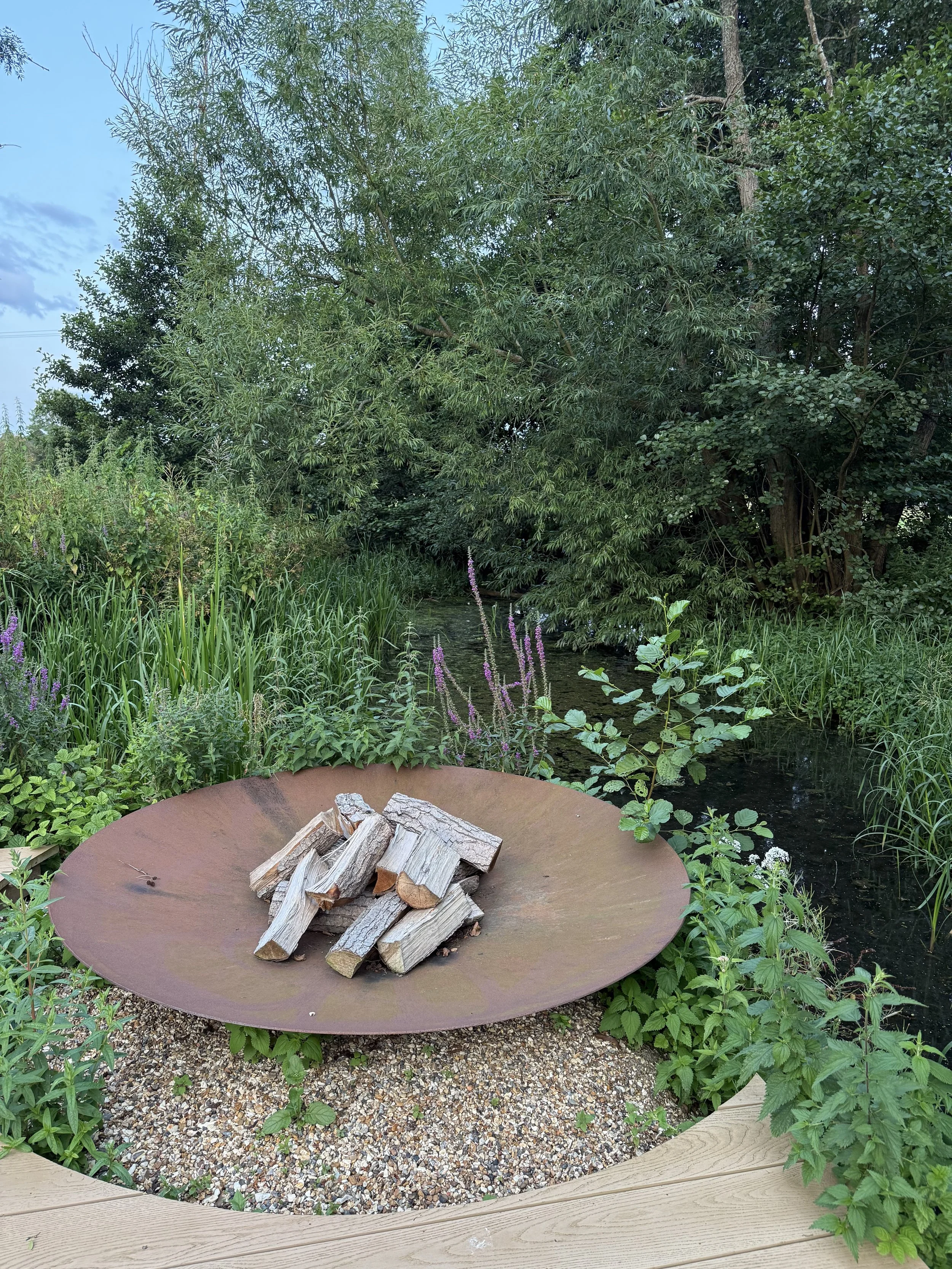 A fire pit with stacked logs, surrounded by lush green plants, tall grasses, and trees, with a small creek visible in the background.