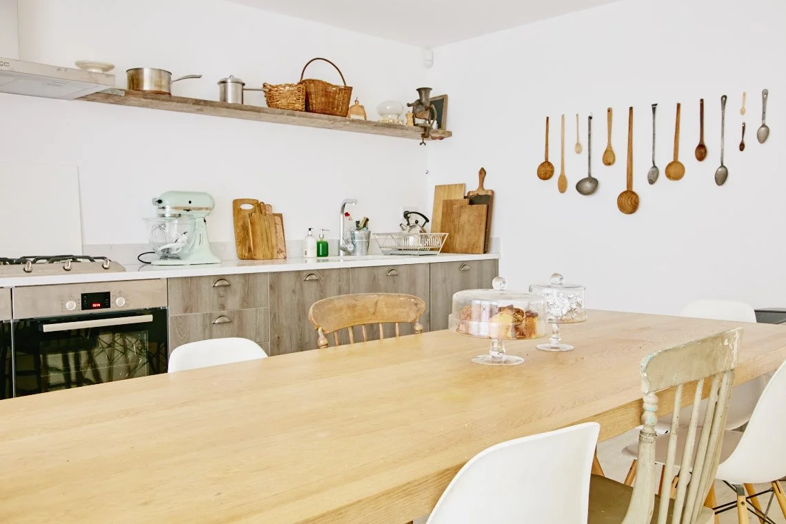 A kitchen with a wooden dining table, vintage and modern chairs, a light-colored countertop, hanging wooden spoons, and shelf with pots, baskets, and kitchen utensils.