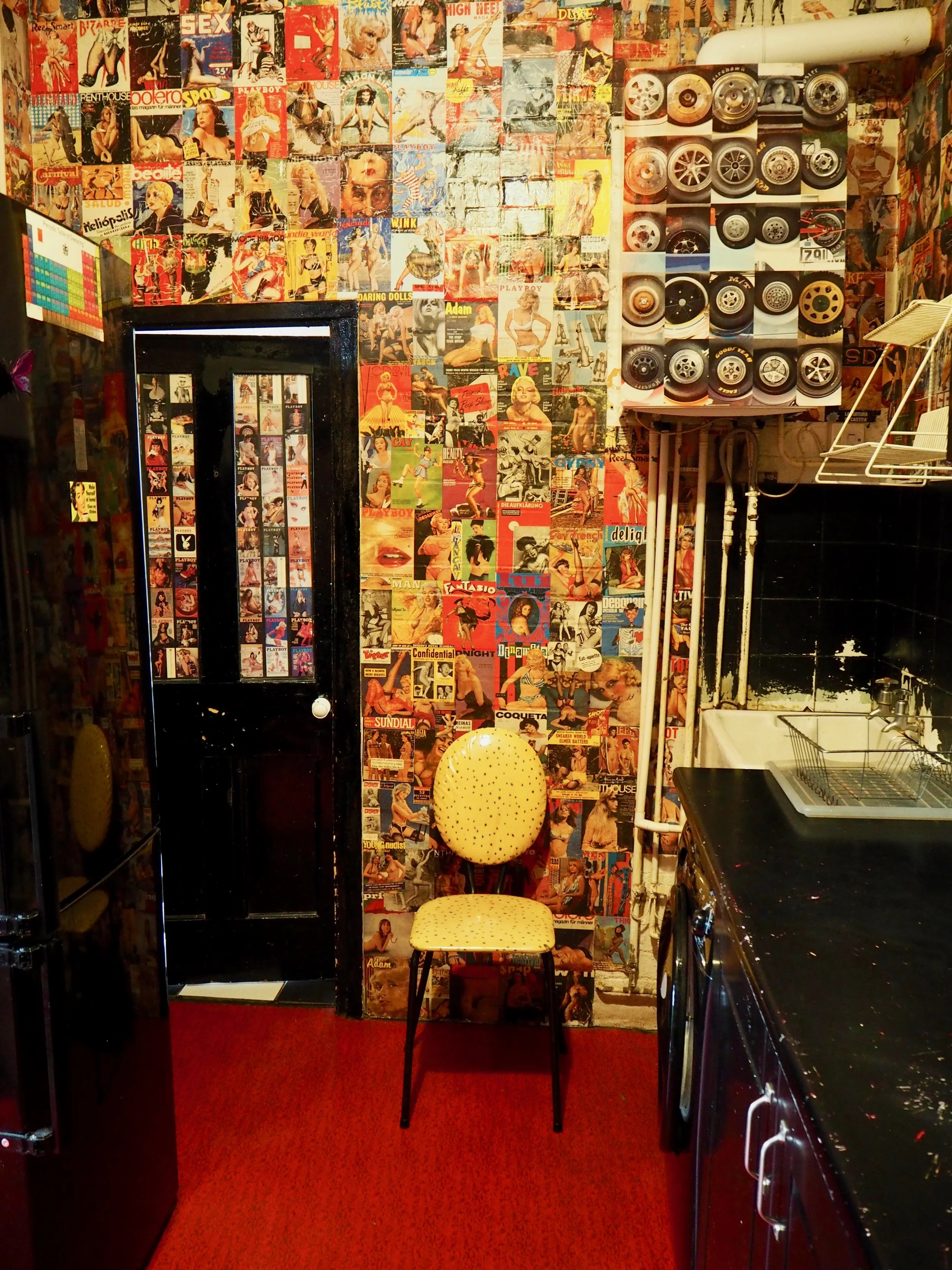 A wall covered with colorful vintage magazine posters, a black door with photo booth strips, a yellow polka dot chair, a collection of wheels on the wall, and part of a black countertop with a sink in a retro-themed room.