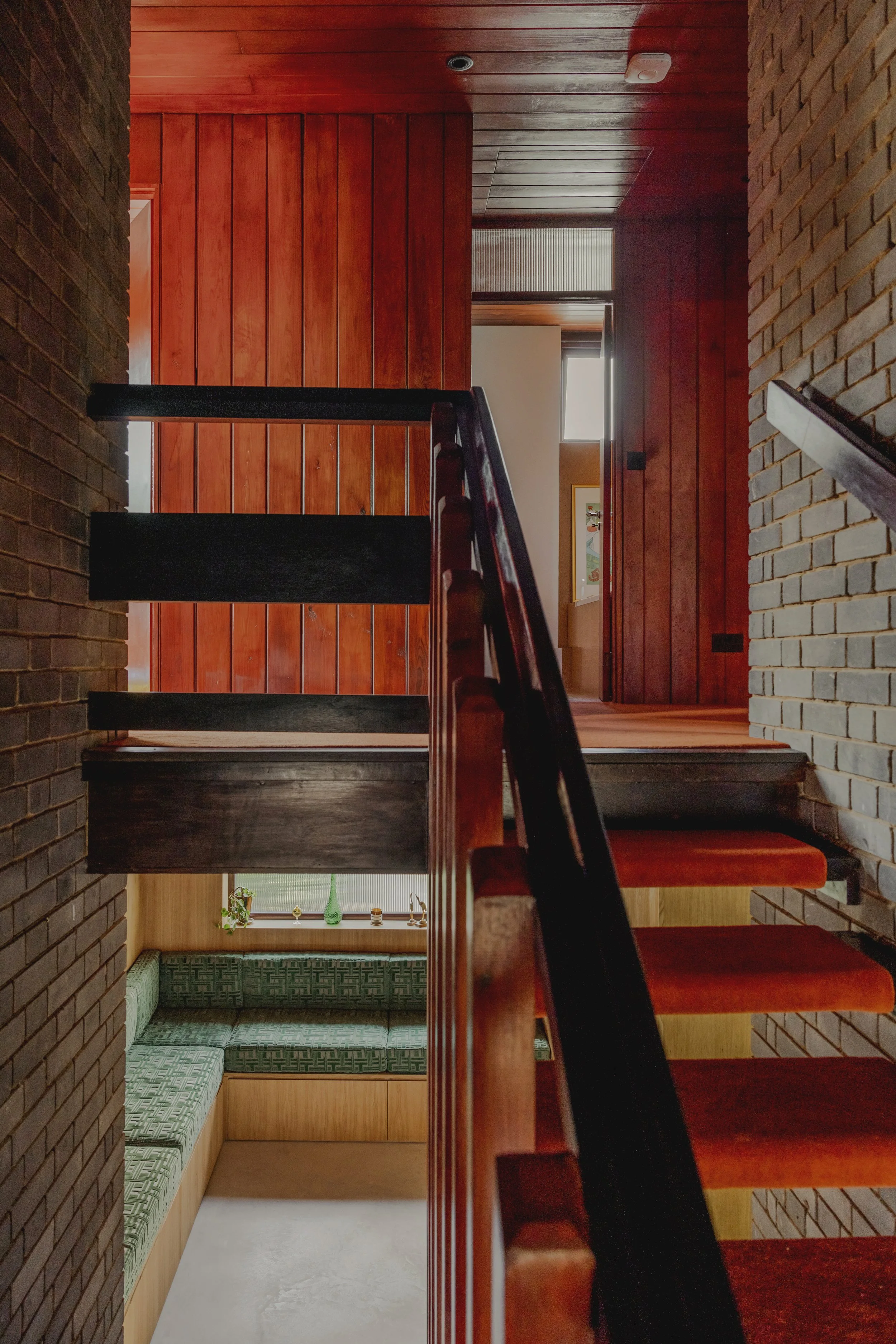 Interior view of a staircase with dark wooden steps, black railing, brick and wood-paneled walls, and a seating nook with green upholstered cushions beneath a window with decorative items.