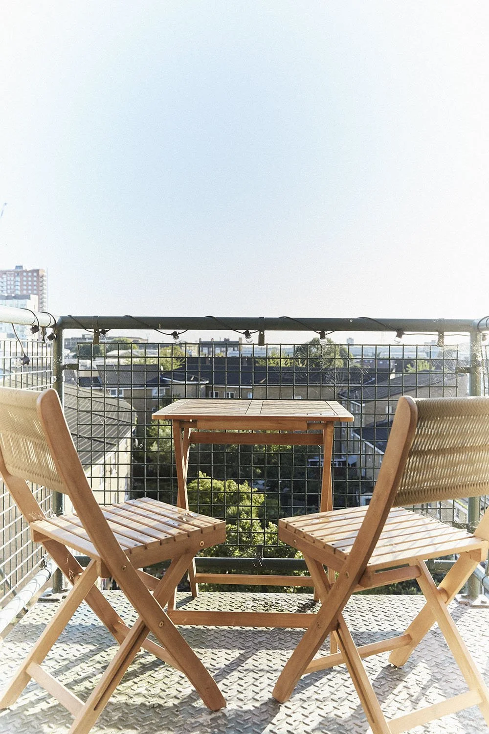 A balcony with two wooden chairs and a small wooden table, overlooking a cityscape with rooftops, trees, and distant buildings under a clear sky.