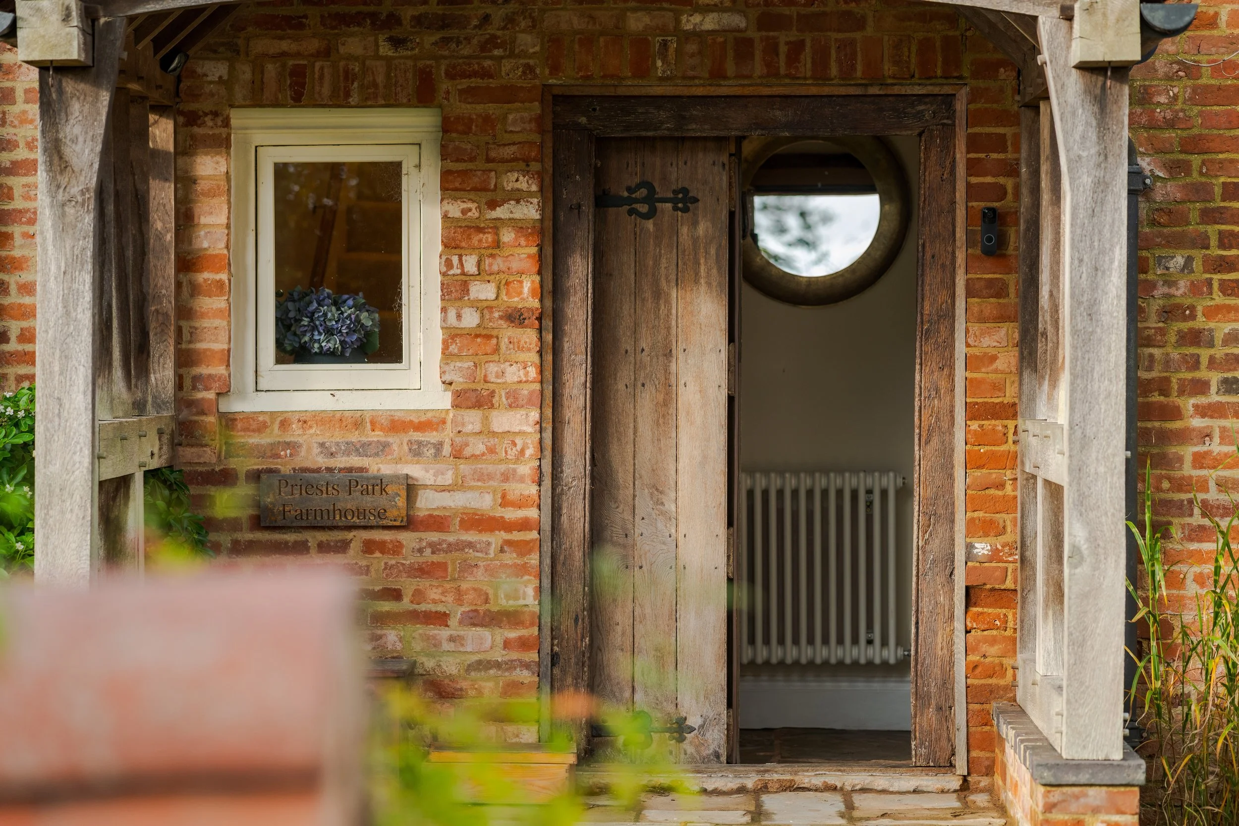 Entrance to Priests Park Farmhouse with a rustic wooden door, a small window with flowers, and brick walls.