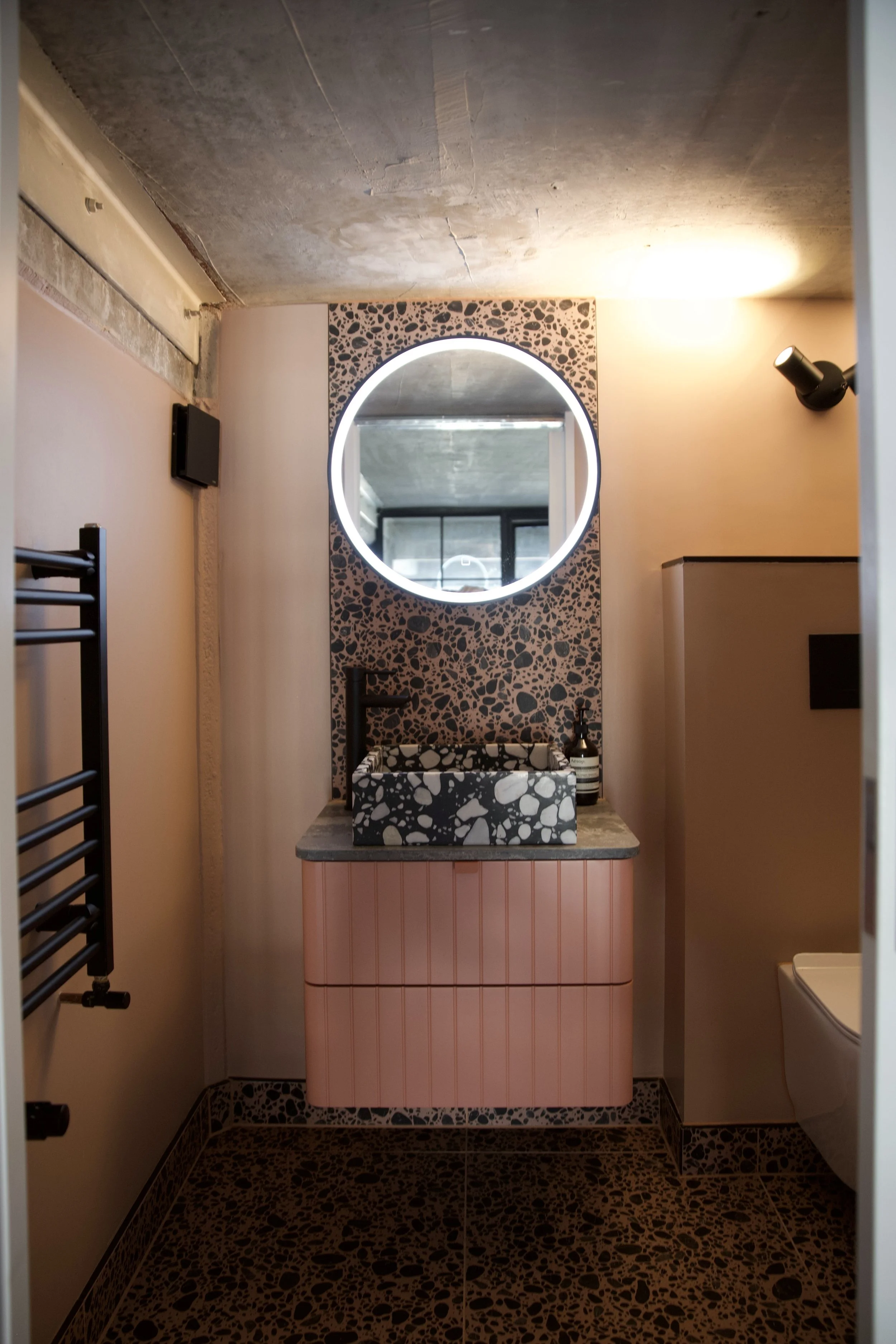 Modern bathroom with a round illuminated mirror, terrazzo sink, pink cabinet, and black fixtures.