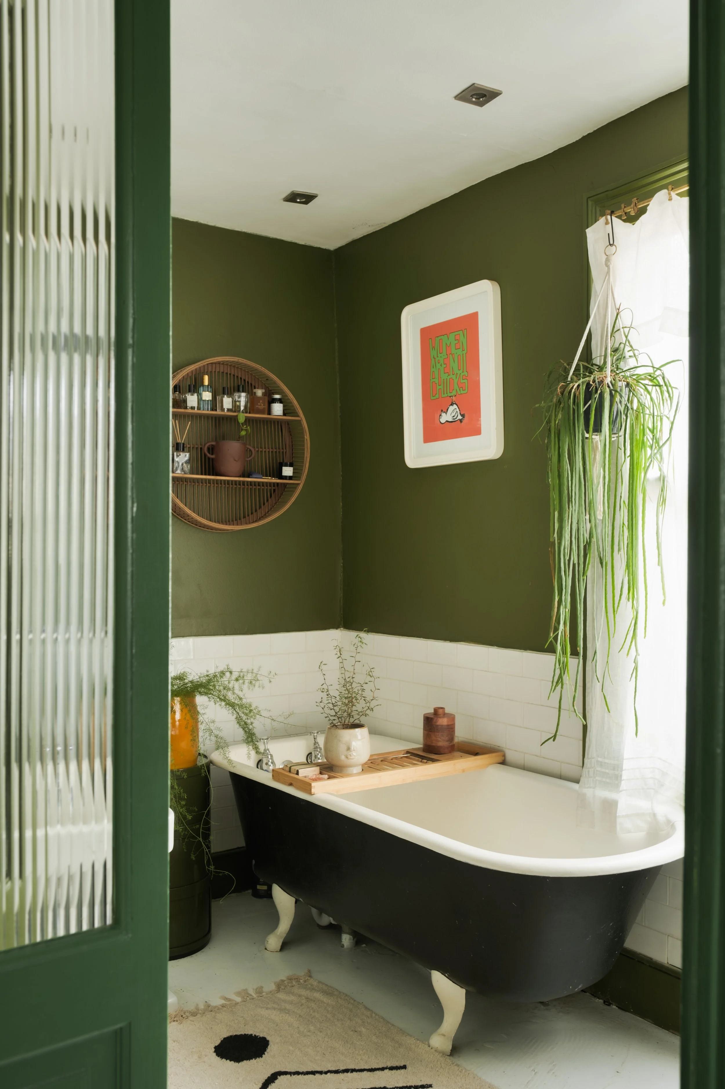 A bathroom with a black clawfoot tub, green walls, hanging plants, framed art, and a white curtain.