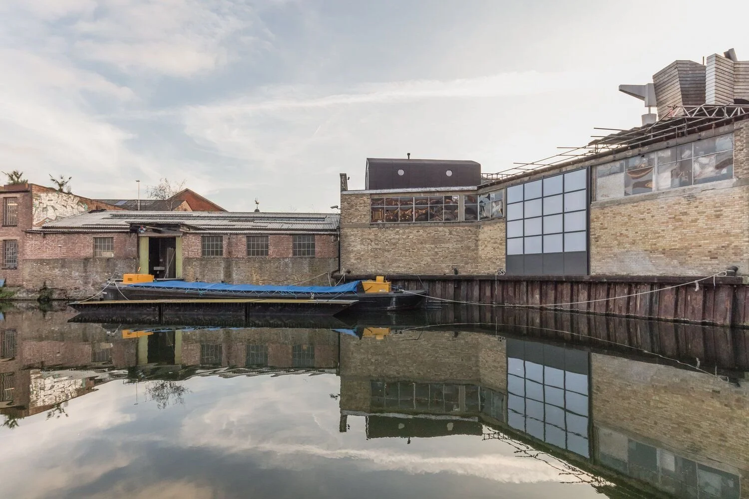 A calm waterway reflecting old brick buildings and modern industrial-style structures with large windows, under a partly cloudy sky.