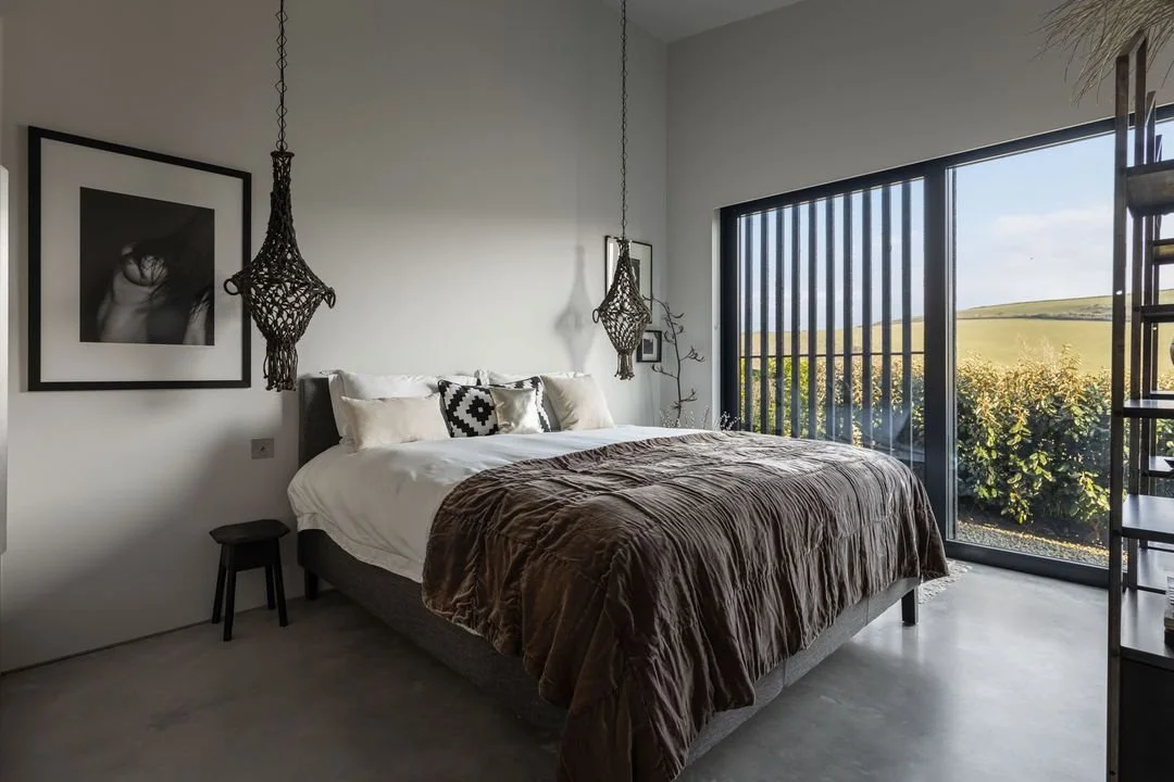 Modern bedroom with large glass window showing a view of greenery and hills, white walls, black framed art, hanging pendant lights, a bed with white and black pillows, and brown bedding.