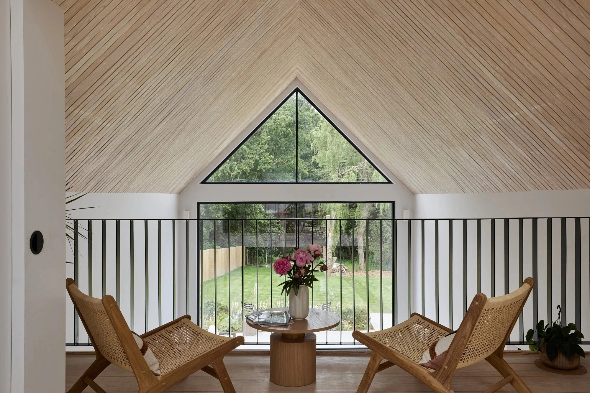 Interior view of a modern attic living space with a wooden ceiling, large black-framed triangular and rectangular windows, a round wooden table with a vase of pink flowers, two wooden chairs with cushions, and a view of a green backyard.